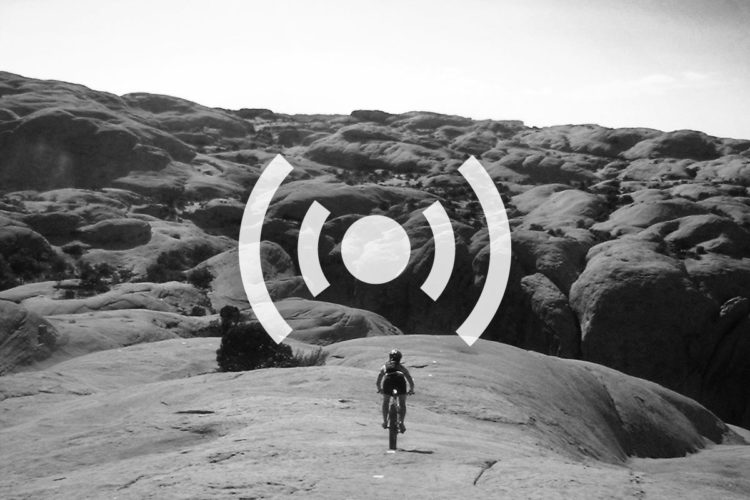 A mountain biker navigating rocky terrain under a clear sky, with rolling hills and scattered vegetation in the background. The image has a black and white filter, and a broadcast symbol overlay is present in the center.