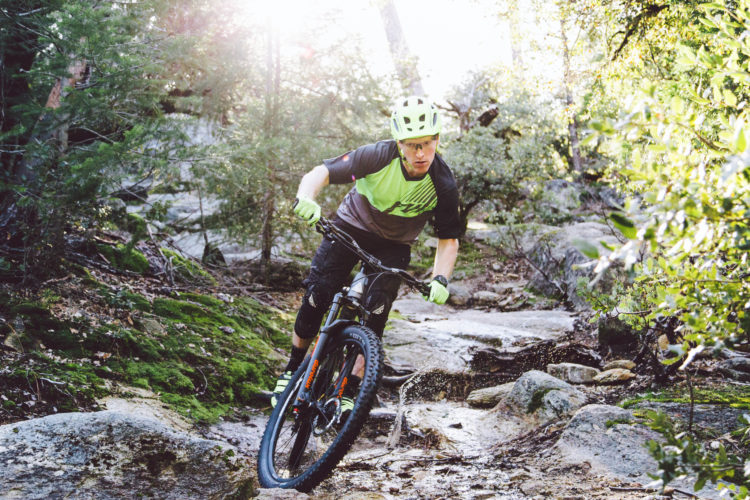 A mountain biker in a bright green and black outfit navigates a rocky trail surrounded by trees. The sun shines through the foliage, creating a dynamic and energetic atmosphere. The biker leans forward for balance as dirt and water spray from the trail, capturing the thrill of off-road cycling.