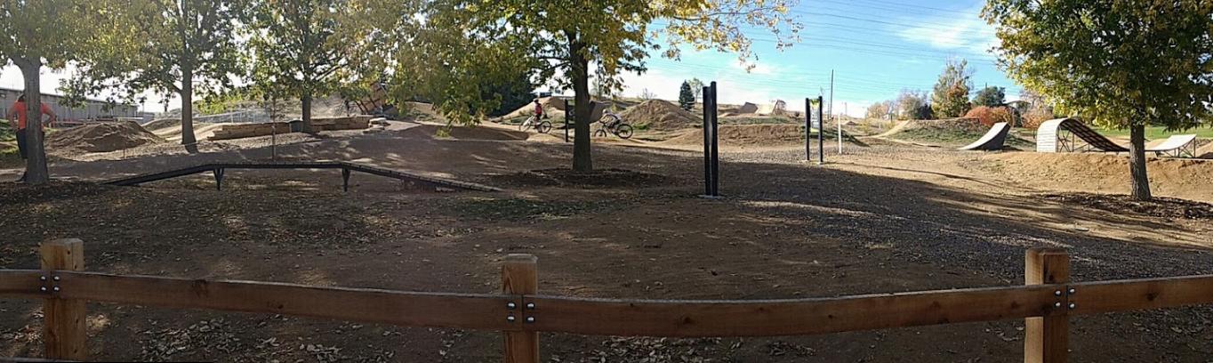 A panoramic view of a bike park featuring dirt ramps and trails surrounded by trees. In the distance, a few cyclists can be seen riding along the dirt paths, while a wooden fence borders the area. The scene is illuminated by sunlight and showcases a clear blue sky with a few clouds. Ruby Hill Park mountain bike trail.
