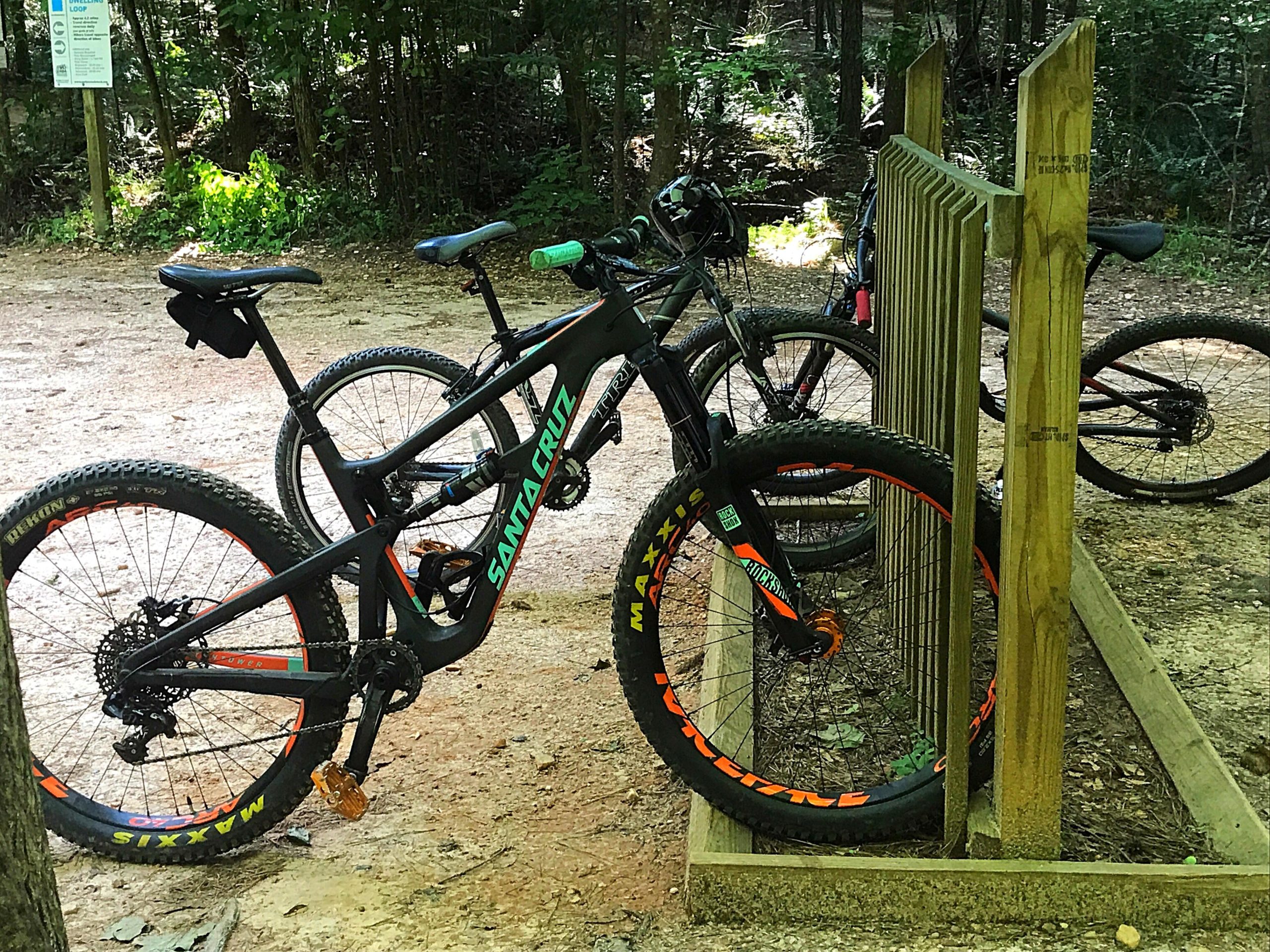 Image of two mountain bikes parked at a wooden bike rack in a forested area. One bike is a black Santa Cruz model with vibrant orange and green accents, while the other bike is partially visible behind it. The background features dense trees and natural surroundings, indicating a trail or outdoor recreational area. Blankets Creek mountain bike trail.