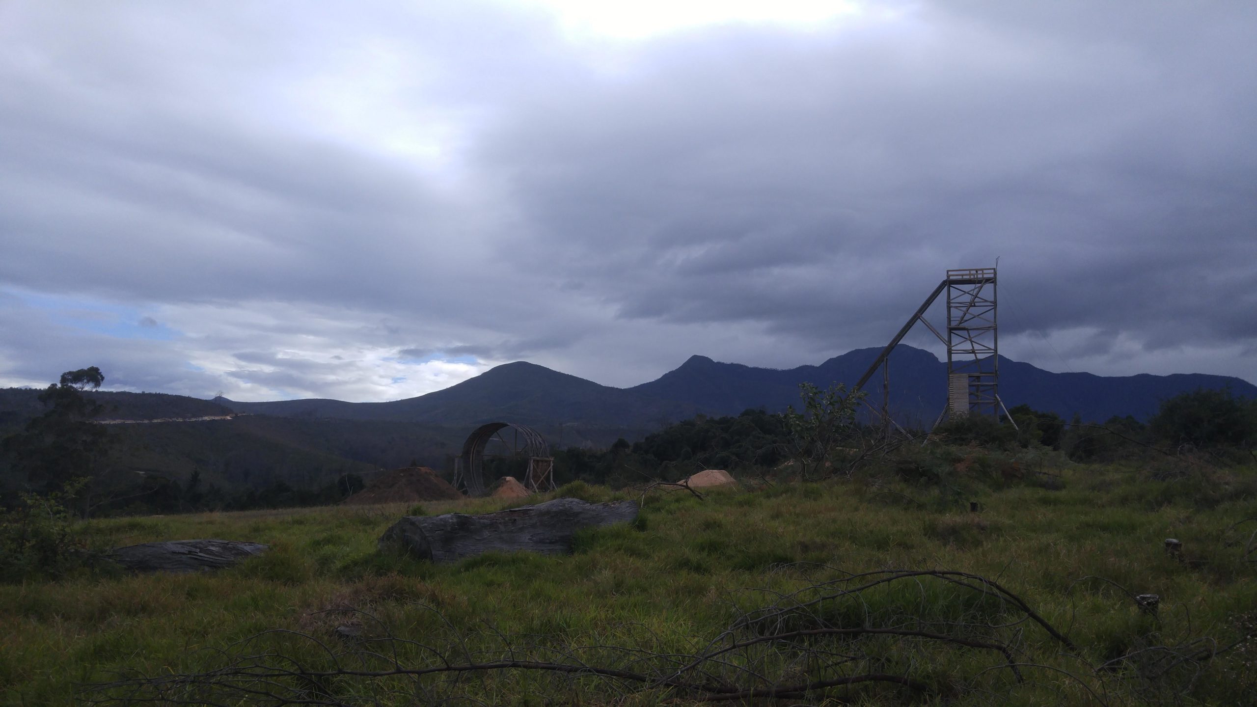 A scenic landscape featuring rolling hills and mountains under a cloudy sky. In the foreground, there is a grassy field with scattered rocks and fallen branches. To the right, a wooden structure resembling a ramp or observation tower stands. A circular wooden frame is partially visible on the left side of the image, indicating possible construction or artistic installation. The overall atmosphere is calm and natural. Garden Route Trail Park mountain bike trail.