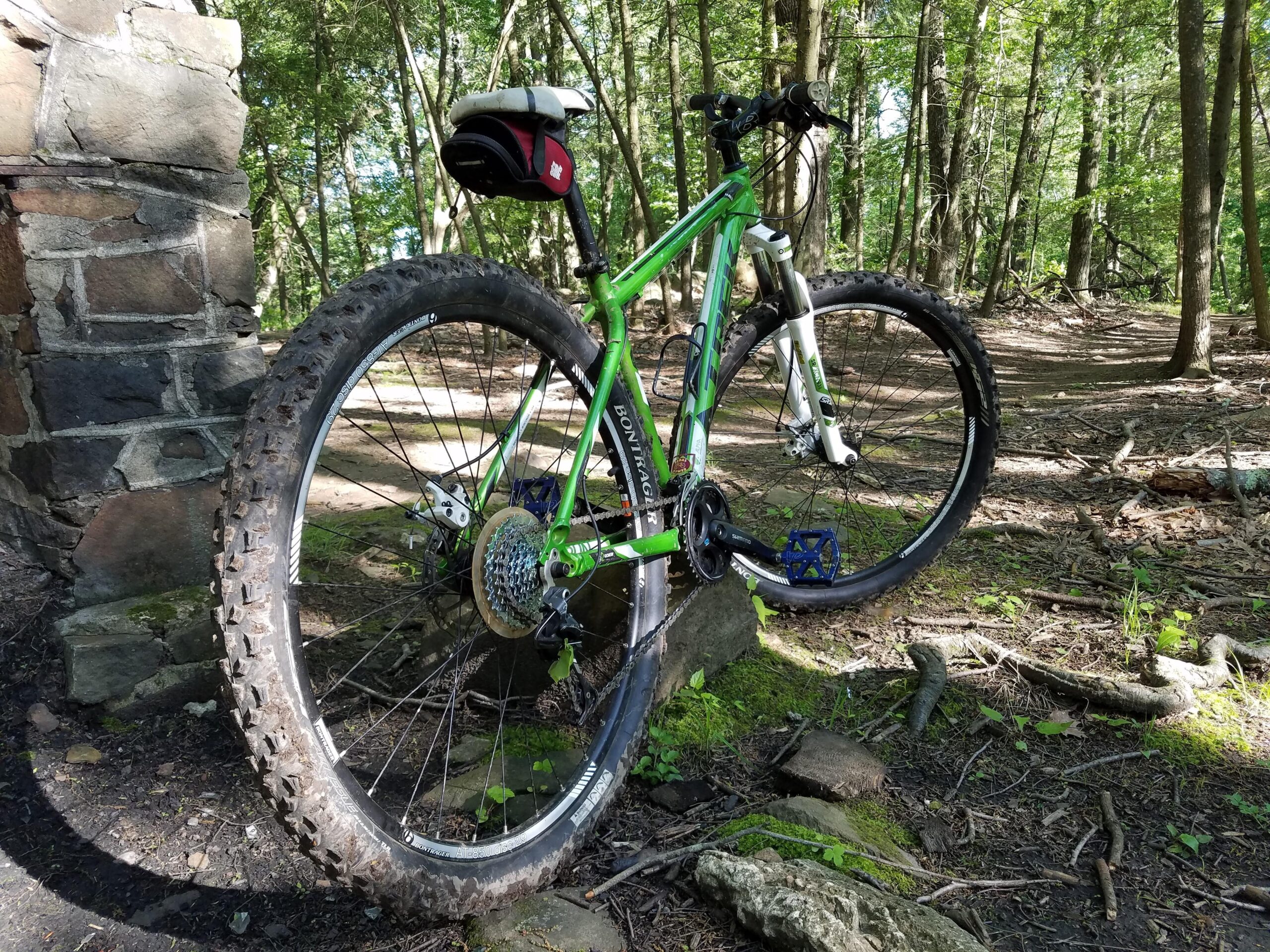Trek Marlin: A green and white mountain bike positioned next to a stone structure in a wooded area, with visible dirt on the tires. The scene features lush greenery and sunlight filtering through the trees, indicating a natural outdoor environment.