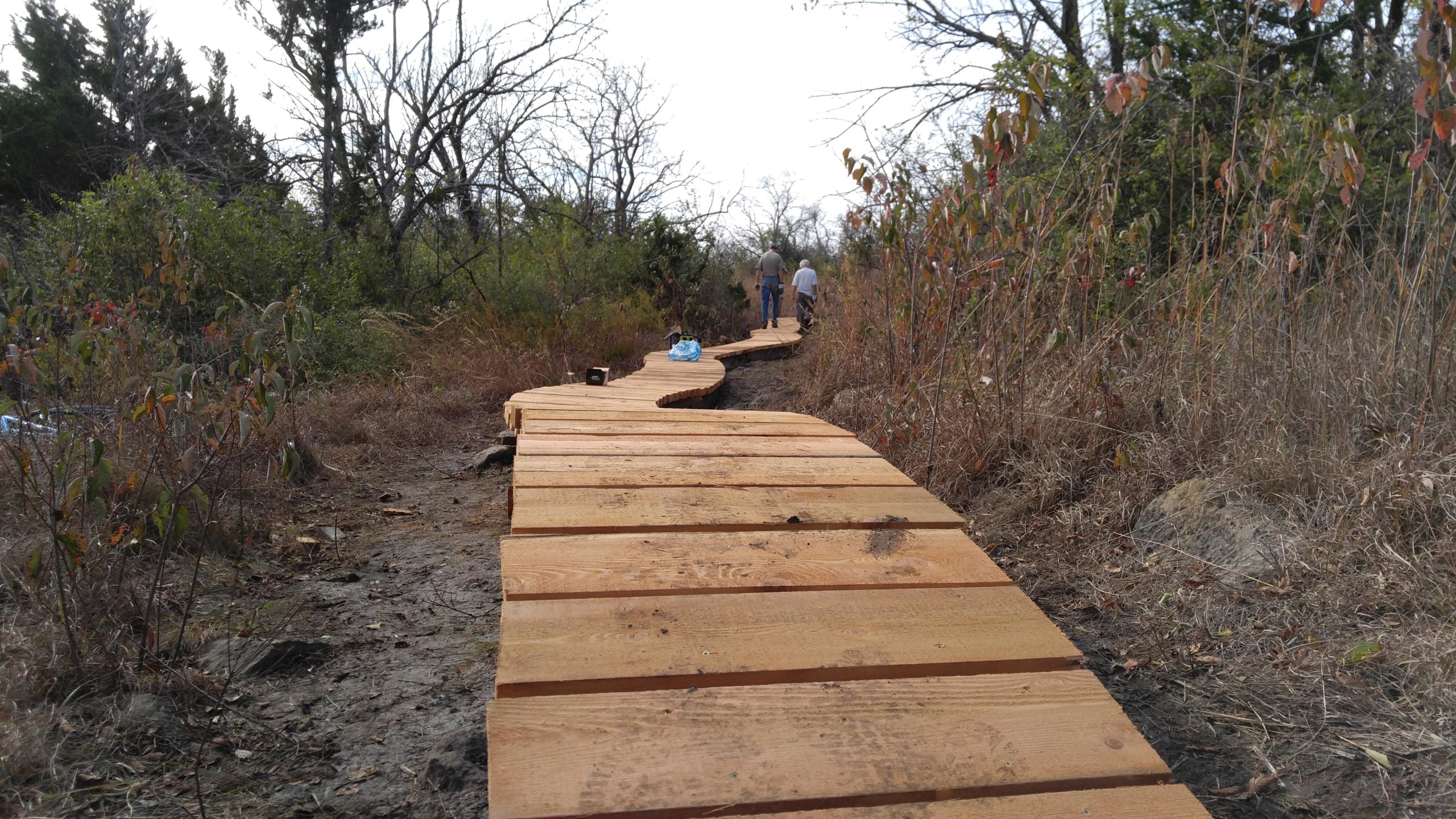 A winding wooden path made of board planks, surrounded by sparse vegetation and trees. Two people are walking along the path in the distance, with tall grass and bushes on either side. The scene is set in an outdoor environment, indicating a trail or walkway through nature. Skyline Park - Burnetts Mound Trail mountain bike trail.