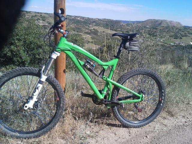 Santa Cruz Nomad: A green mountain bike leaning against a wooden post, set against a scenic landscape with rolling hills and a clear blue sky. The bike features rugged tires and a small black saddle bag attached.