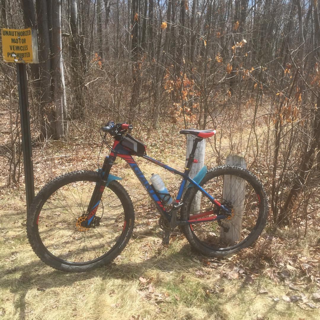 Giant Talon 29er: A mountain bike resting on a grassy area beside a trail with a sign that reads "Unauthorized Motor Vehicles Prohibited." The surroundings feature bare trees and remnants of fall leaves, indicating early spring. The bike is equipped with a water bottle and a small bag attached to the frame.