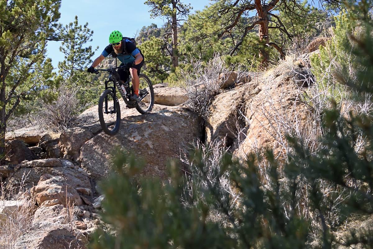 Niner RIP 9: A mountain biker navigating rocky terrain on a sunny day, surrounded by trees and shrubs. The rider wears a bright green helmet and a blue shirt, demonstrating skill and balance as they maneuver over the rocks.