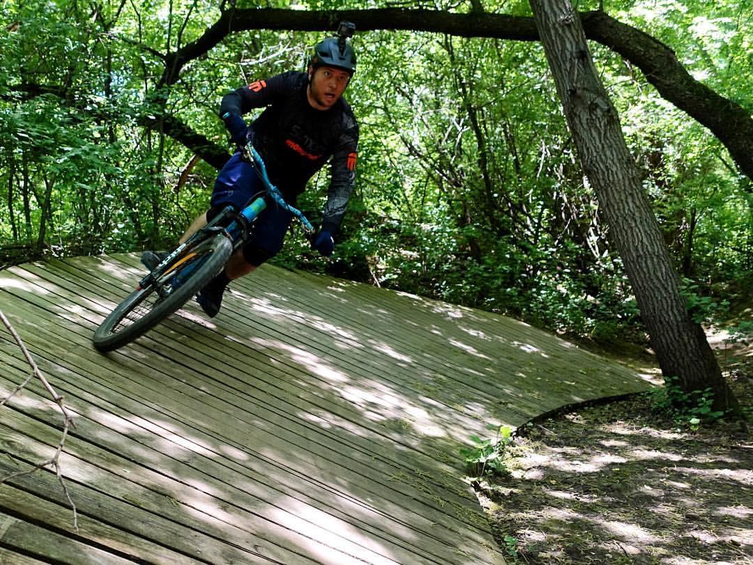 A mountain biker wearing a helmet and protective gear navigates a wooden ramp in a lush, green forest. The biker is leaning into the turn, demonstrating skill and balance while riding the trail. Sunlight filters through the trees, creating a vibrant, natural backdrop. Quarry Ridge Park mountain bike trail.