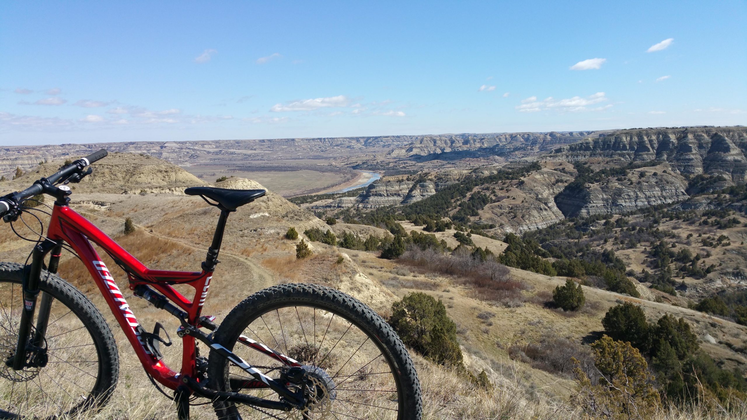 A red mountain bike is positioned on a hillside overlooking a vast canyon landscape with rolling hills and a winding river below. The sky is clear with a few clouds, showcasing a bright blue backdrop. The scene captures the essence of outdoor adventure and natural beauty. Maah Daah Hey mountain bike trail.