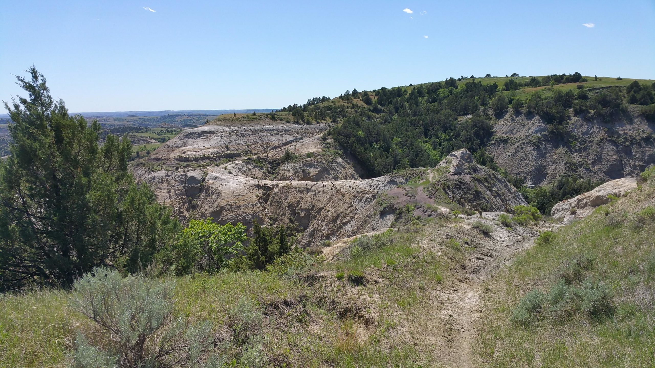 A scenic view of a rugged landscape featuring steep rock formations and rolling hills under a bright blue sky. Lush greenery and shrubs are present in the foreground, while distant hills rise in the background, creating a picturesque natural setting. Maah Daah Hey mountain bike trail.