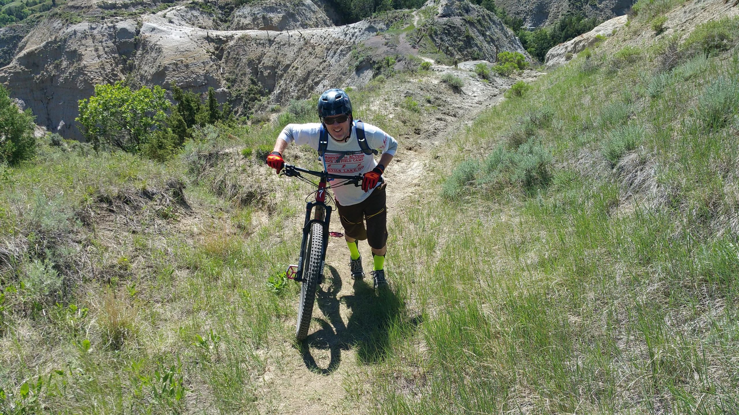 A mountain biker pushing their bike up a grassy trail, surrounded by hilly terrain and trees in the background. The rider is wearing a helmet, sunglasses, and gloves, with bright yellow socks and shorts, smiling as they navigate the incline on a sunny day. Maah Daah Hey mountain bike trail.
