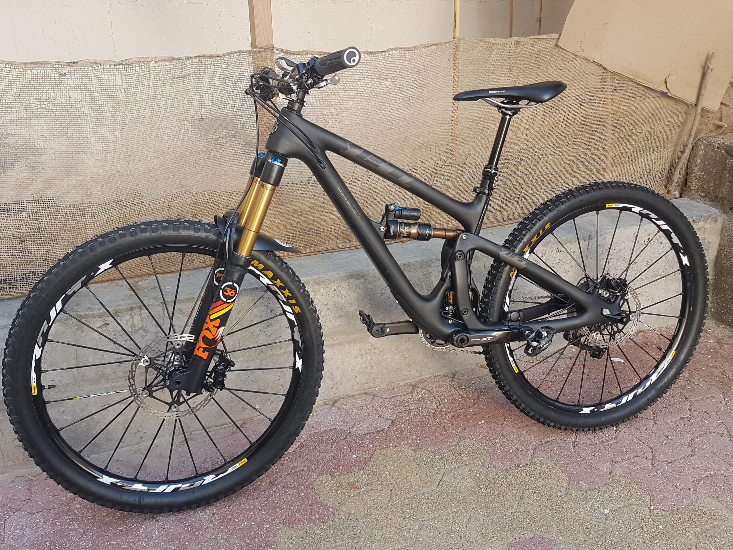 Yeti SB6c Switch Infinity: A mountain bike with a sleek black frame and golden front suspension fork, positioned against a textured background. The bike features large tires with visible tread patterns, a dropper seat post, and disc brakes. It's resting on a patterned surface, hinting at an outdoor setting.