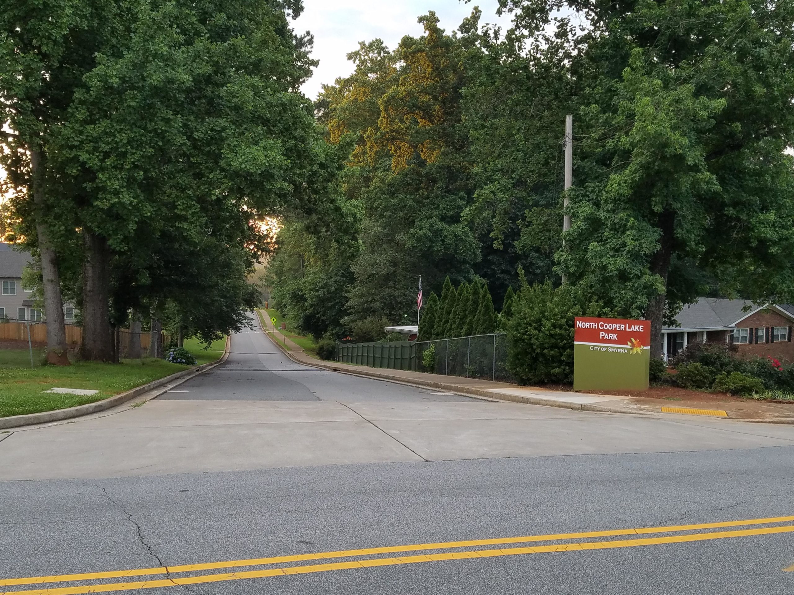 A view of a peaceful road leading to North Cooper Lake Park, surrounded by lush trees and greenery, with a sign indicating the park's entrance. Residential homes line the left side, and the road gently slopes upwards in the distance. The scene is illuminated by soft, natural light, suggesting early morning or late afternoon. North Cooper Lake Park Trail mountain bike trail.
