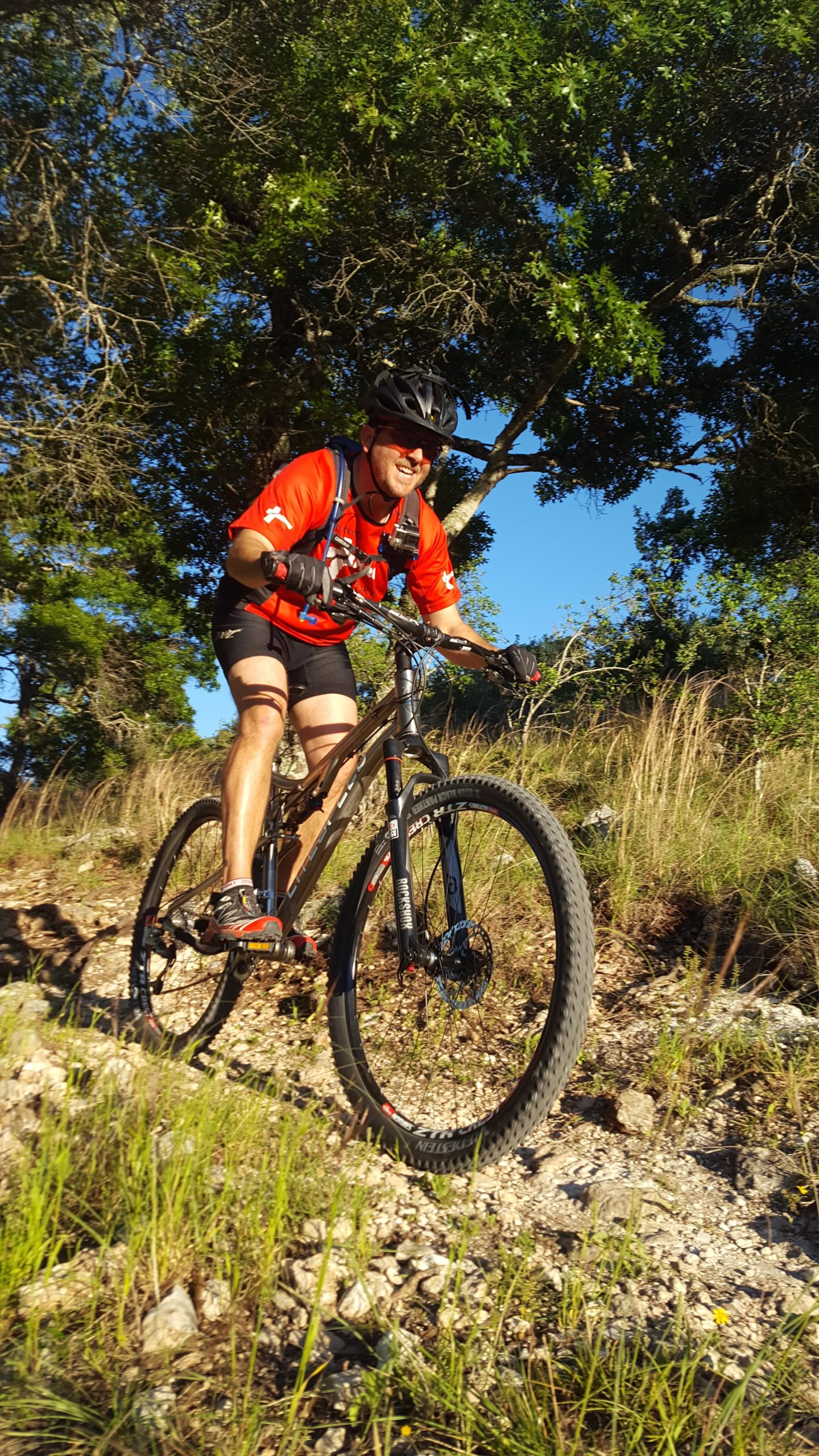 A mountain biker in a red shirt and black shorts rides downhill on a rocky trail surrounded by greenery. The sun casts a warm light, highlighting the determination on the rider's face as he navigates the terrain. Flat Rock Ranch mountain bike trail.