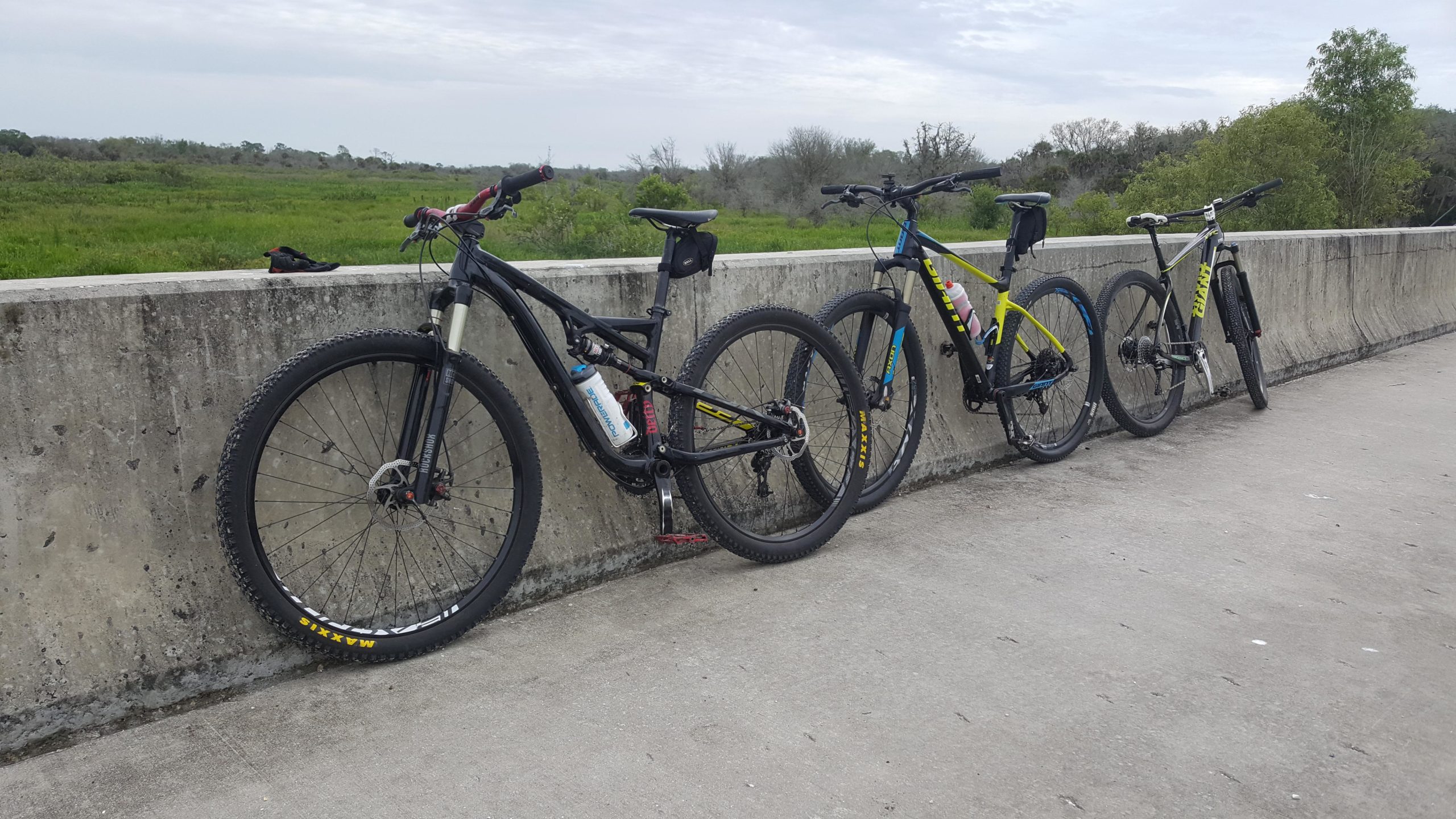 Three mountain bikes are leaning against a concrete barrier, with a grassy landscape and cloudy sky in the background. The bikes vary in color, including black, yellow, and blue accents, and feature distinct tires designed for off-road use. The setting suggests a trail or biking path. BoldlyGo mountain bike trail.