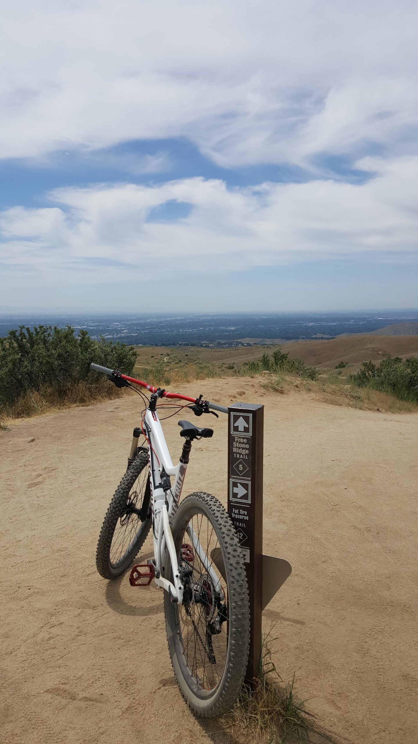 A mountain bike leaning against a trail marker on a dirt path, with a scenic view of rolling hills and a blue sky partially covered by clouds in the background. The trail sign points to different paths, indicating the Free Stone Ridge Trail and Fat Tire Traverse Trail. Kestral, Crestline, Sidewinder, Fat Tire Traverse and Freestone mountain bike trail.