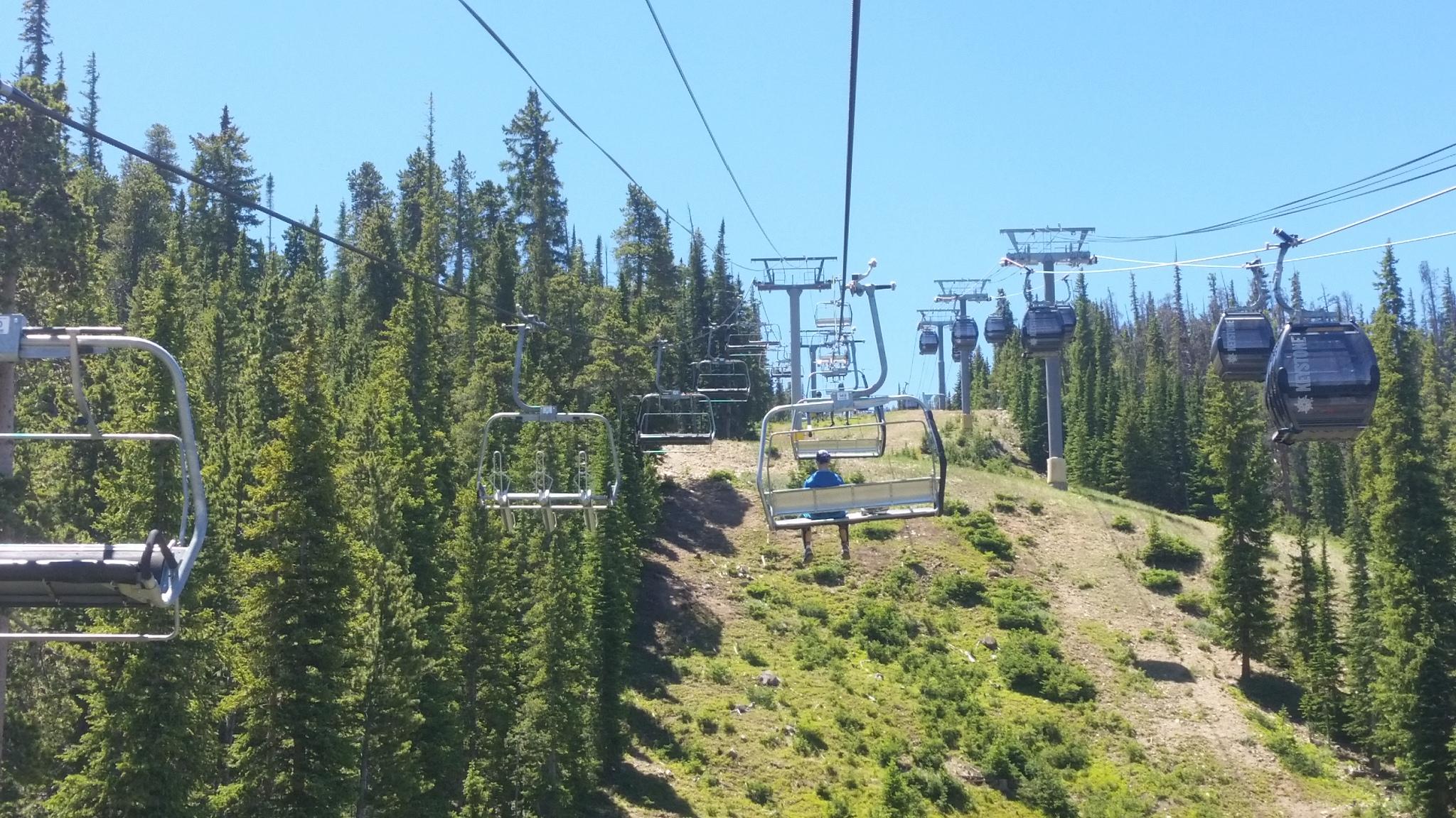 A scenic view of a chairlift and gondola system in a mountainous area, surrounded by tall green pine trees and clear blue skies. The image captures multiple chairlifts ascending a slope, with one person seated on a lift and distant gondolas visible in the background. Keystone Resort Bike Park mountain bike trail.
