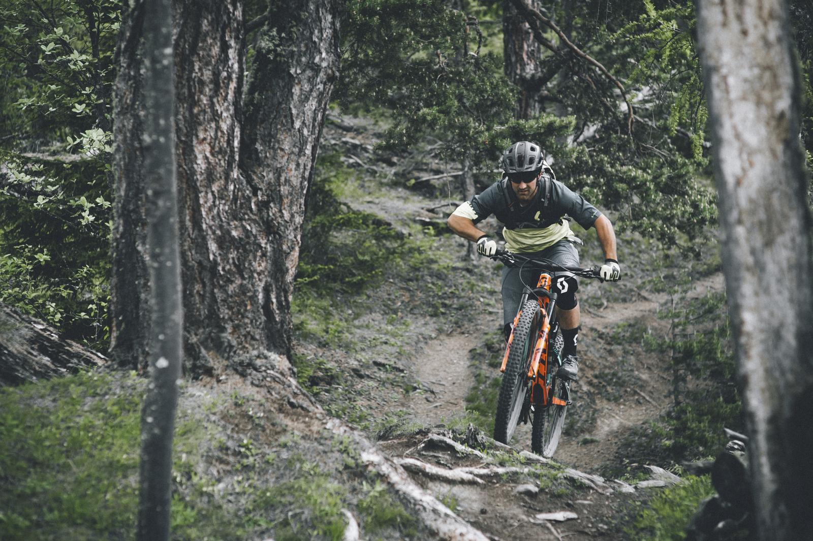 Scott Genius: A mountain biker navigating a rugged, forested trail, focused and in motion, surrounded by trees and rocks. The cyclist is wearing protective gear and riding an orange mountain bike, showcasing an adventurous outdoor atmosphere.