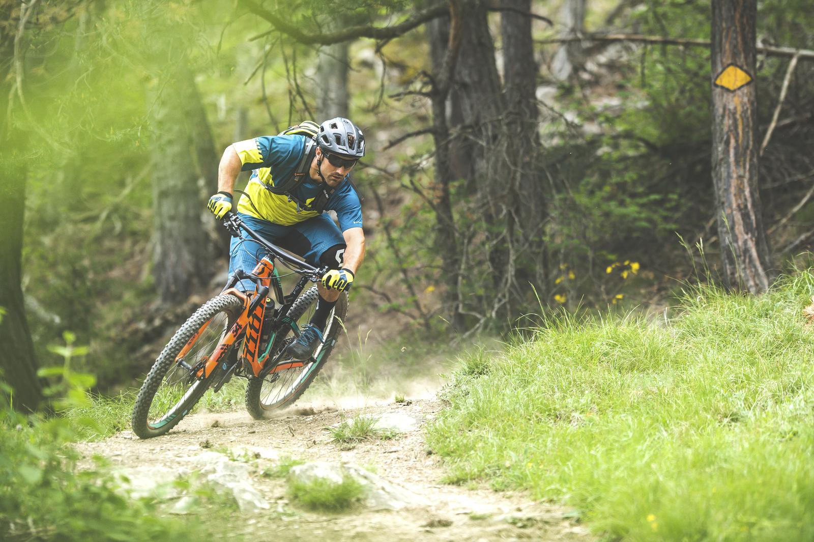 Scott Genius: A mountain biker in a blue and yellow jersey leans into a turn on a dirt trail surrounded by trees, creating a cloud of dust. The cyclist wears a helmet and gloves, showcasing an active and adventurous moment in outdoor sports.