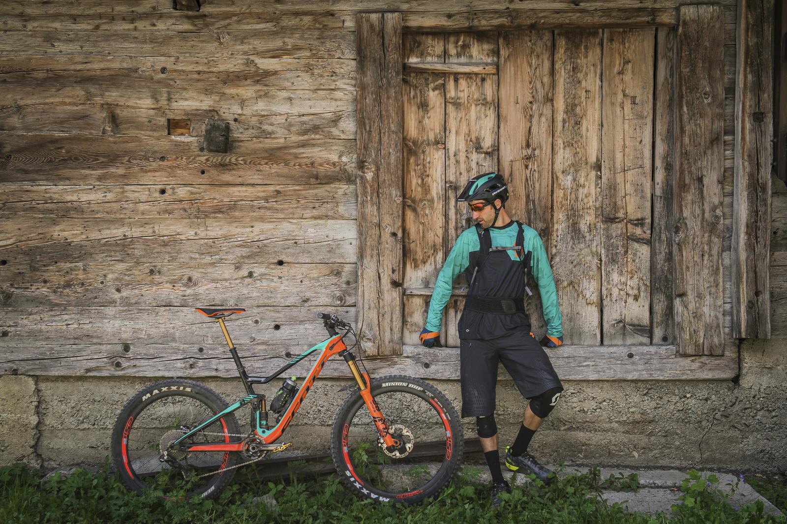 Scott Genius: A mountain biker wearing a teal and black outfit, helmet, and sunglasses leans casually against a rustic wooden wall next to his mountain bike, which has an orange and teal color scheme. The scene is set in a natural outdoor environment with green grass in the foreground.
