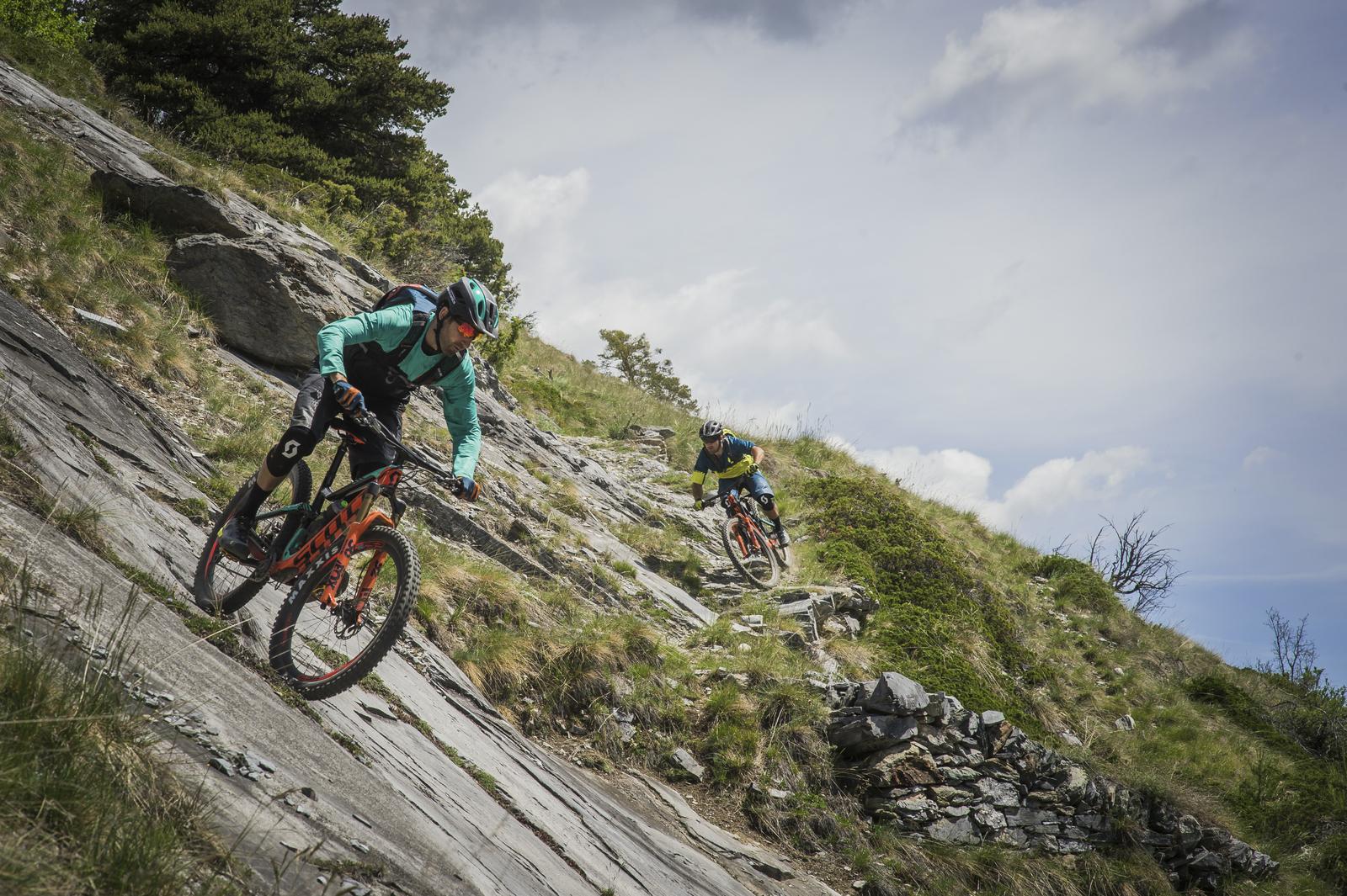 Scott Genius: Two mountain bikers navigating a rocky, sloped terrain in a scenic outdoor setting. One rider, wearing a teal jacket and helmet, leans into the slope while the second rider, dressed in a yellow and black outfit, follows closely behind. The landscape features green grass and scattered rocks under a partly cloudy sky.
