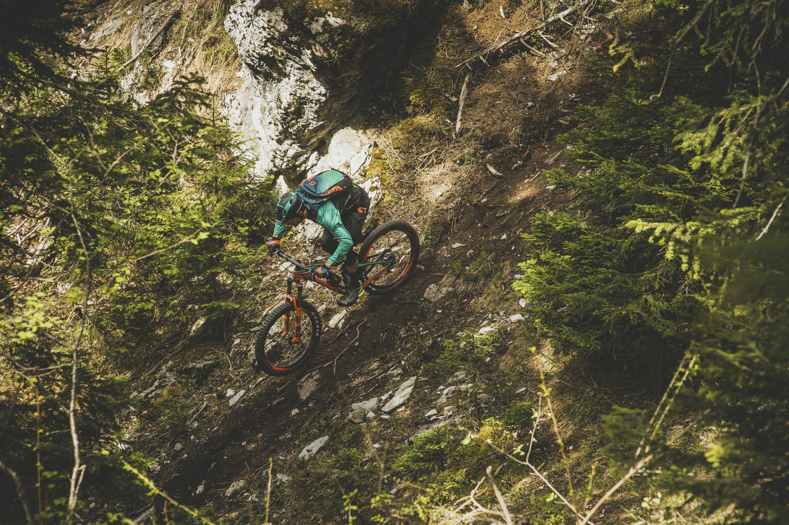 Scott Genius: A mountain biker navigates a rocky downhill trail surrounded by dense greenery and rocky terrain. The biker is dressed in a teal outfit and helmet, demonstrating skill and agility as they ride down the slope. Sunlight filters through the trees, highlighting the natural environment.