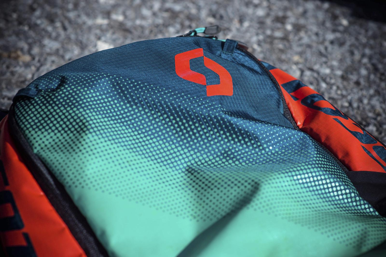 Scott Genius: Close-up of a multi-colored backpack featuring a gradient design with a pattern of dots. The fabric has a dark blue upper section, transitioning to green, and includes a prominent red logo. The background shows a textured surface, suggesting an outdoor setting.
