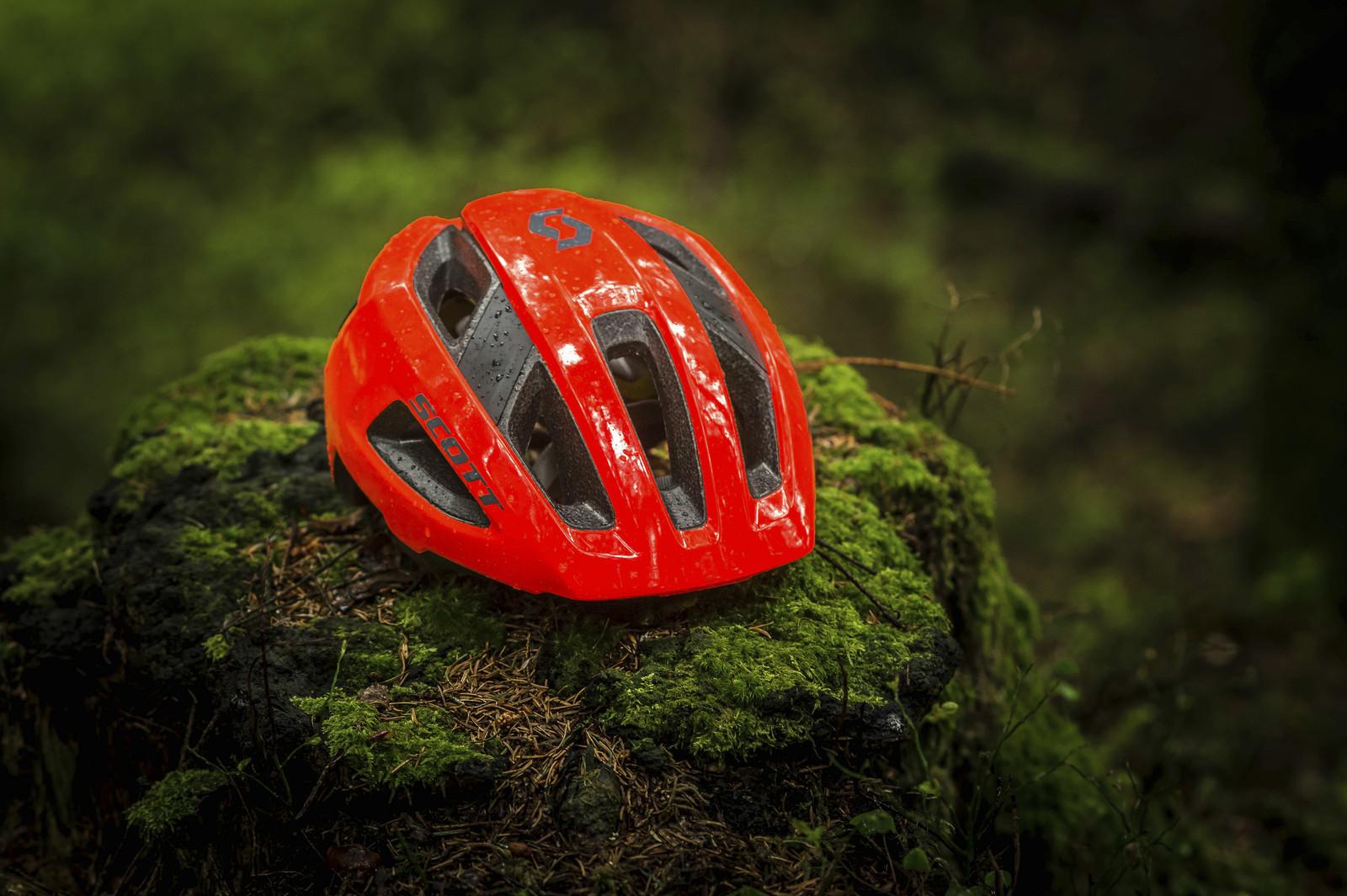 Scott Genius: A bright red bicycle helmet resting on a moss-covered rock in a lush, green forest setting, with droplets of water on its surface, indicating recent rain.