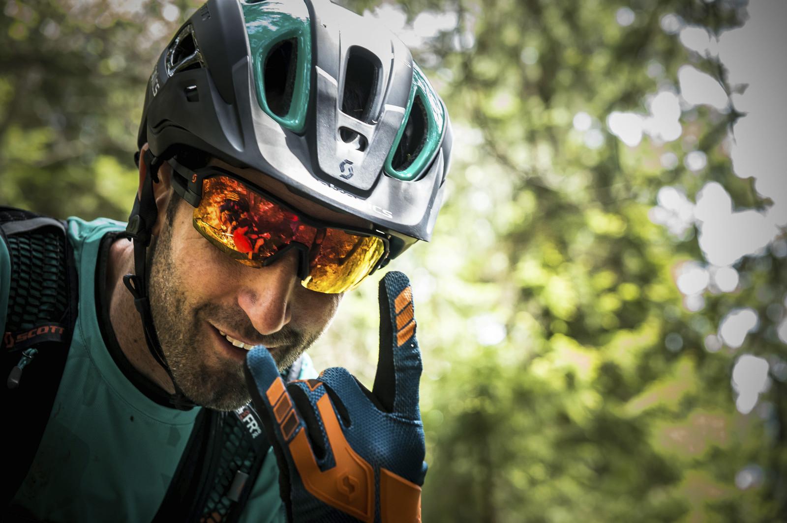 Scott Genius: A mountain biker wearing a helmet and sunglasses smiles and makes a hand gesture in a forested area, with dappled sunlight filtering through the trees in the background.