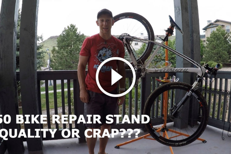 A person standing on a balcony next to a bike repair stand with a bicycle mounted on it. The background includes trees and buildings. Text overlay asks if the $50 bike repair stand is quality or not, with a play button icon in the center.