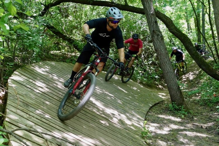 A group of four mountain bikers navigating a wooden trail in a lush green forest. The first rider, wearing a black shirt and a helmet, is leaning into a curve, while the others follow on parallel paths. Sunlight filters through the trees, casting dappled shadows on the ground.
