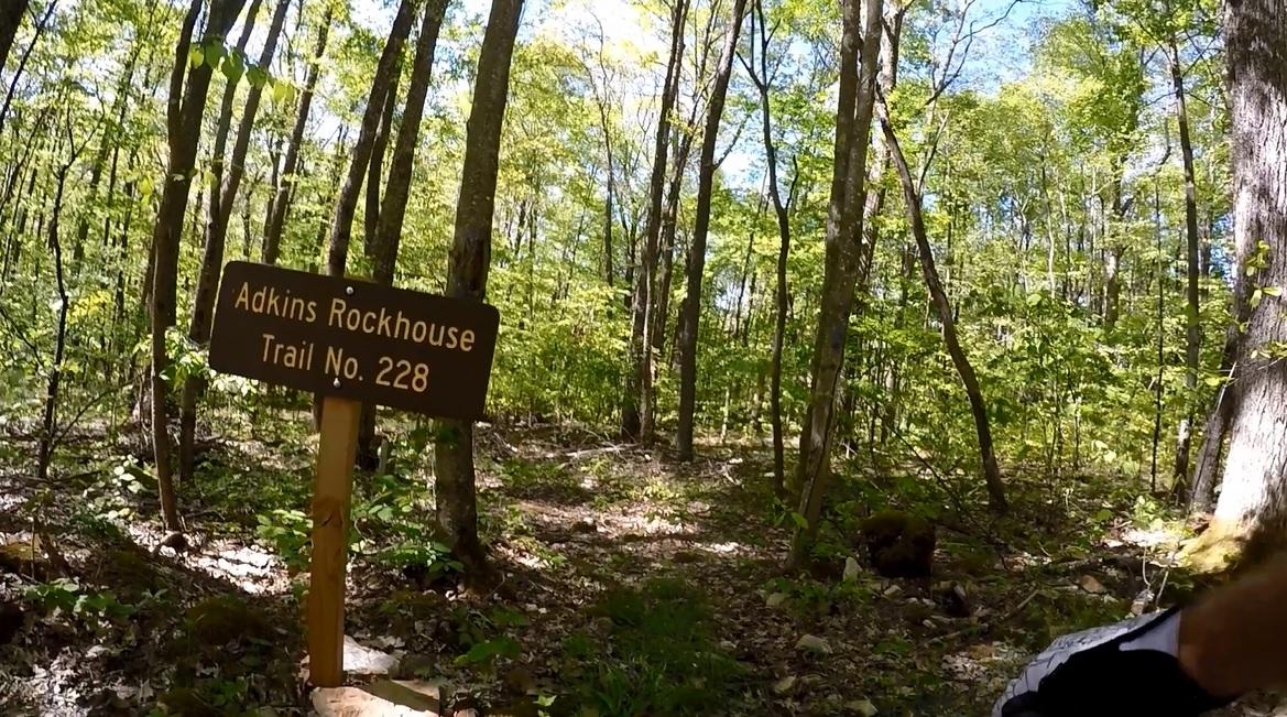 A wooden sign indicating "Adkins Rockhouse Trail No. 228" in a lush green forest with tall trees and sunlight filtering through the leaves. Adkins Rockhouse mountain bike trail.