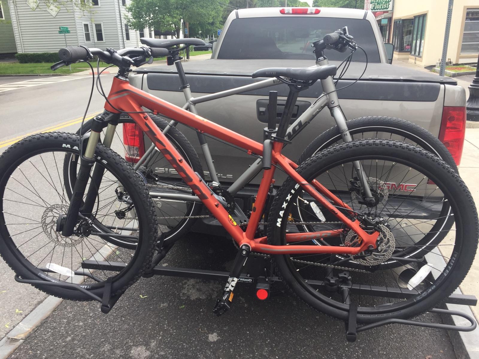 Kona Cinder Cone: Two mountain bikes are secured on a hitch-mounted bike rack attached to the back of a pickup truck. The bike in the foreground is orange and features a suspension fork, while the bike behind it is gray. The truck's tailgate is partially visible, along with a glimpse of the street and nearby buildings in the background.