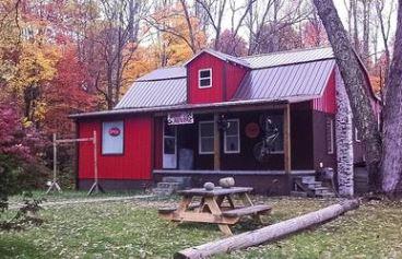 A charming red cabin with a metal roof, set in a wooded area adorned with autumn foliage. The front yard features a picnic table and a log, while the cabin showcases multiple windows and a welcoming entrance.