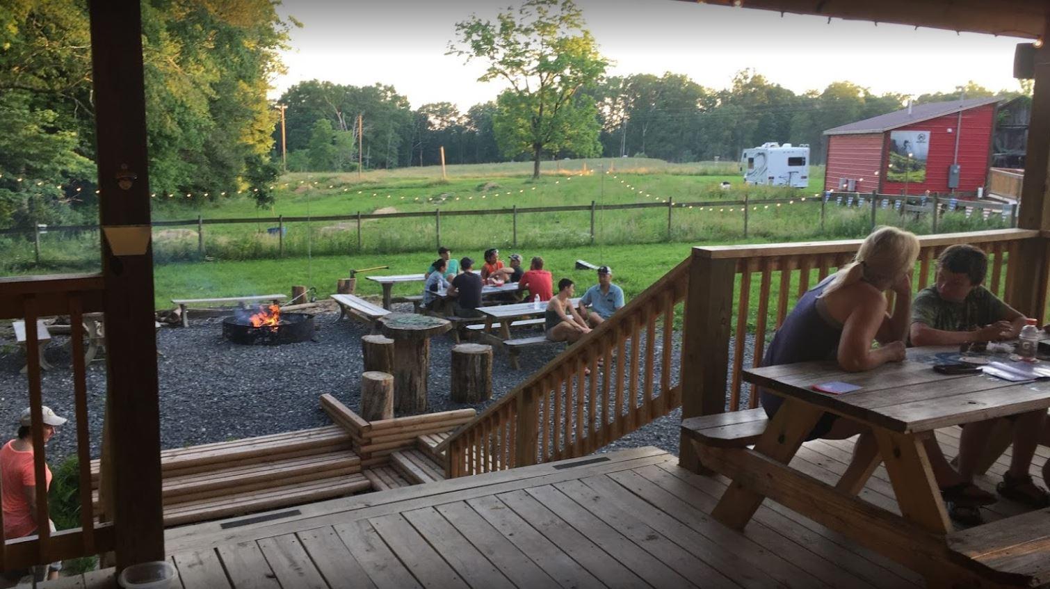 A scenic outdoor area featuring a wooden deck overlooking a fire pit surrounded by seating. In the background, groups of people enjoy each other's company around picnic tables, with lush greenery and a barn visible in the distance. Soft evening lighting adds to the relaxed atmosphere of the gathering.