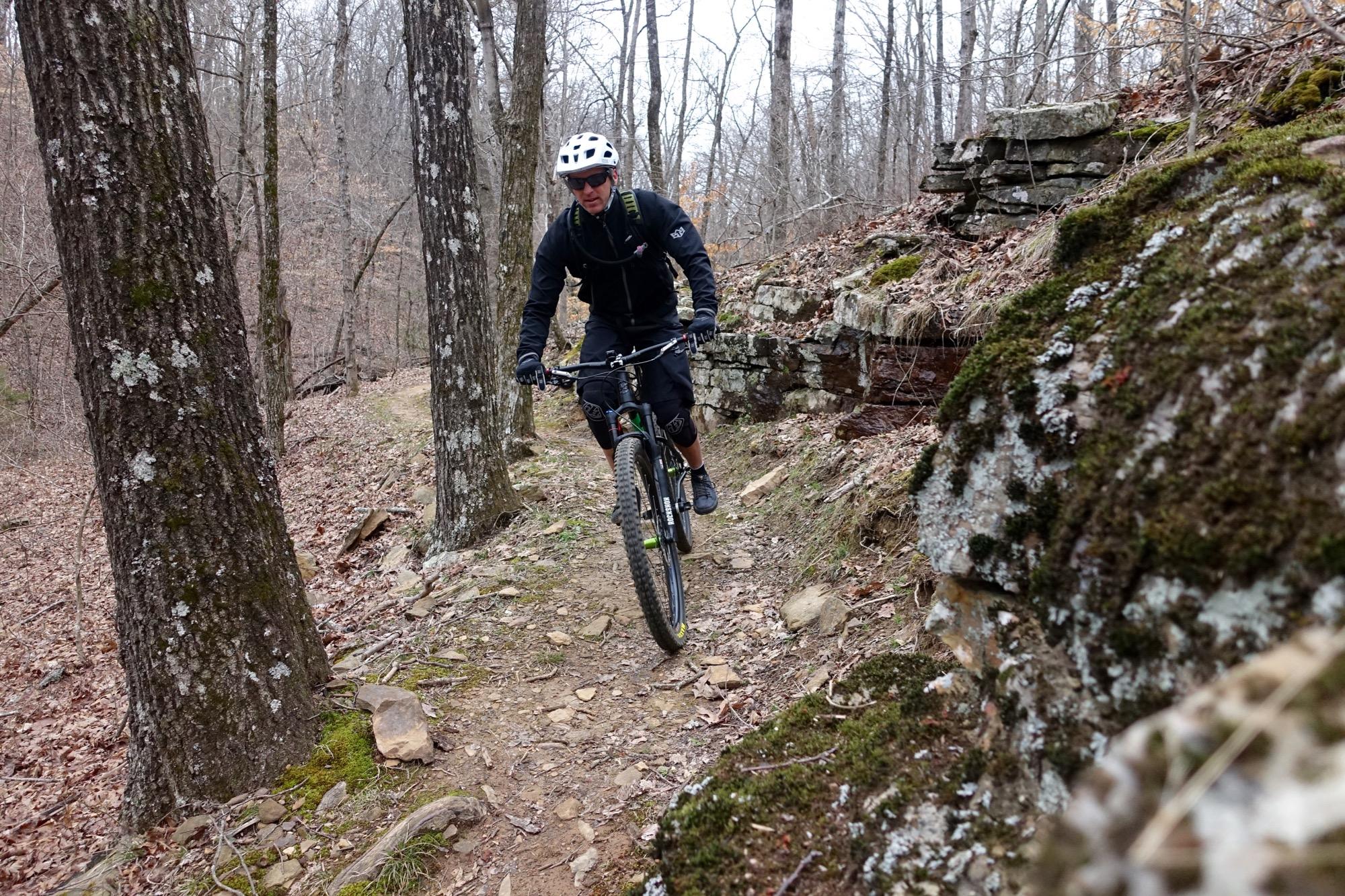 A mountain biker navigates a narrow dirt trail surrounded by trees and rocky terrain in a wooded area. The cyclist is wearing a helmet, sunglasses, and biking gear, and is focused on maneuvering through the natural landscape. Upper Buffalo Headwaters Trail System mountain bike trail.