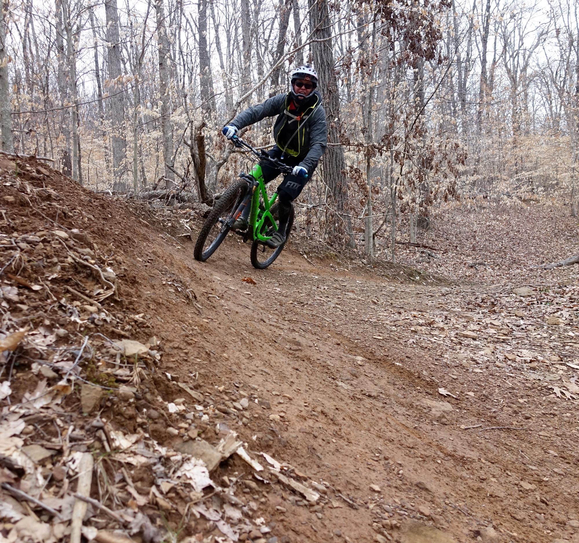 A mountain biker in a gray jacket and helmet rides a green bike down a dirt trail surrounded by bare trees and fallen leaves. The rider leans into a turn, showcasing a dynamic action shot in a natural setting during autumn. Upper Buffalo Headwaters Trail System mountain bike trail.