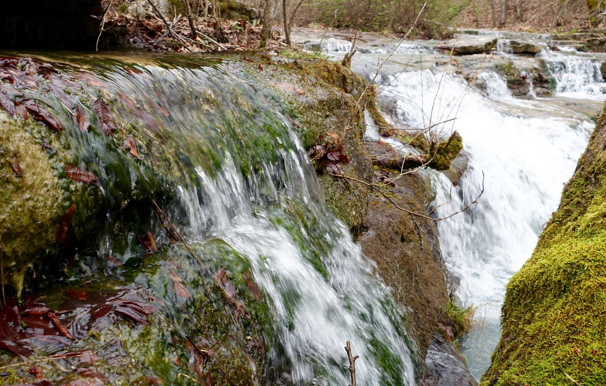 A close-up view of a small waterfall flowing over moss-covered rocks, surrounded by fallen leaves and bare branches. The water cascades gently, creating a serene and natural landscape with a soft, blurred background of additional water flow and greenery. Upper Buffalo Headwaters Trail System mountain bike trail.