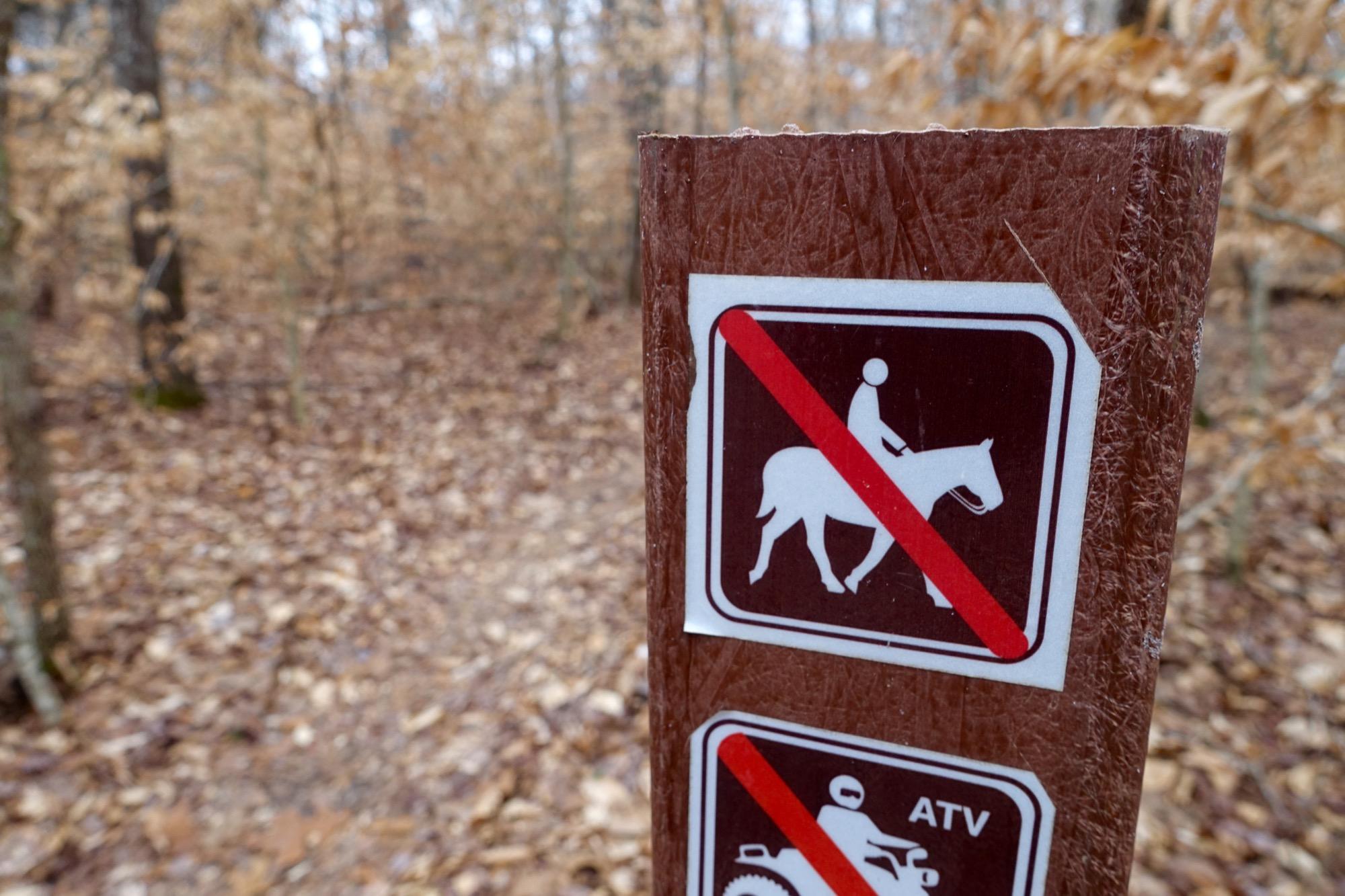 A wooden sign in a wooded area displaying prohibition symbols for horseback riding and ATVs, with a background of fallen leaves and trees. Upper Buffalo Headwaters Trail System mountain bike trail.