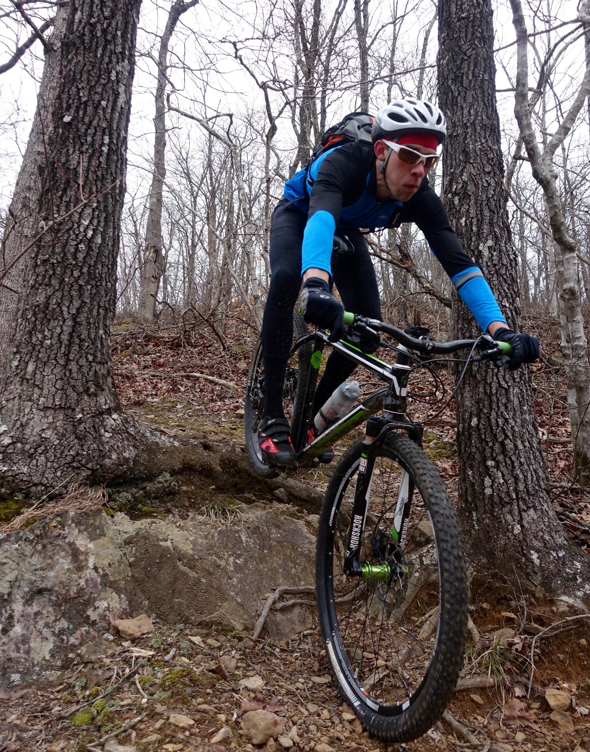 A mountain biker in a blue and black outfit is jumping over a rocky terrain in a forested area, with bare trees in the background. The rider wears a helmet and sunglasses, showcasing an intense focus as they navigate the trail. Upper Buffalo Headwaters Trail System mountain bike trail.