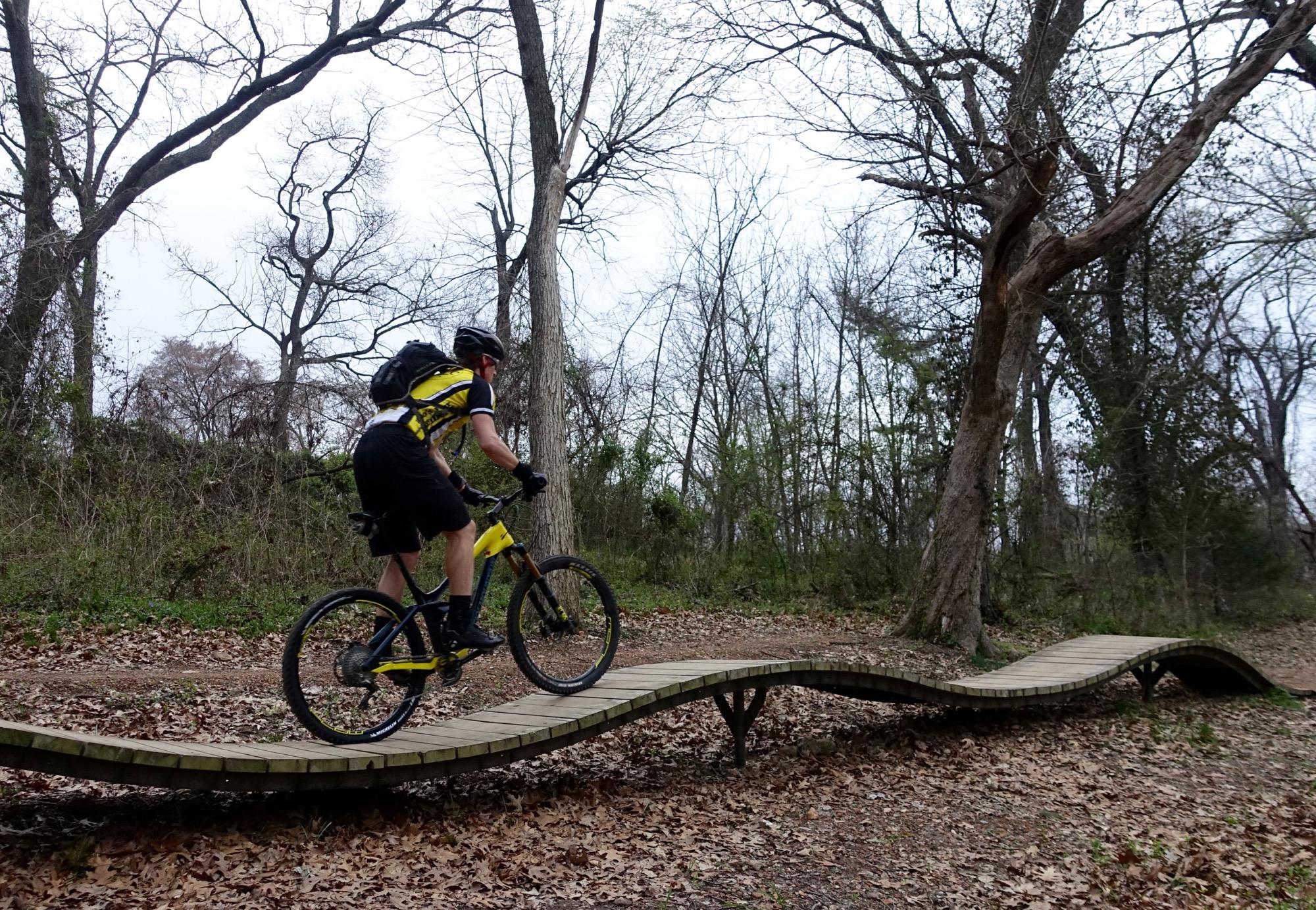 A mountain biker in yellow and black gear riding on a wooden bridge through a wooded area, surrounded by bare trees and fallen leaves, on a cloudy day. Slaughter Pen Trail mountain bike trail.