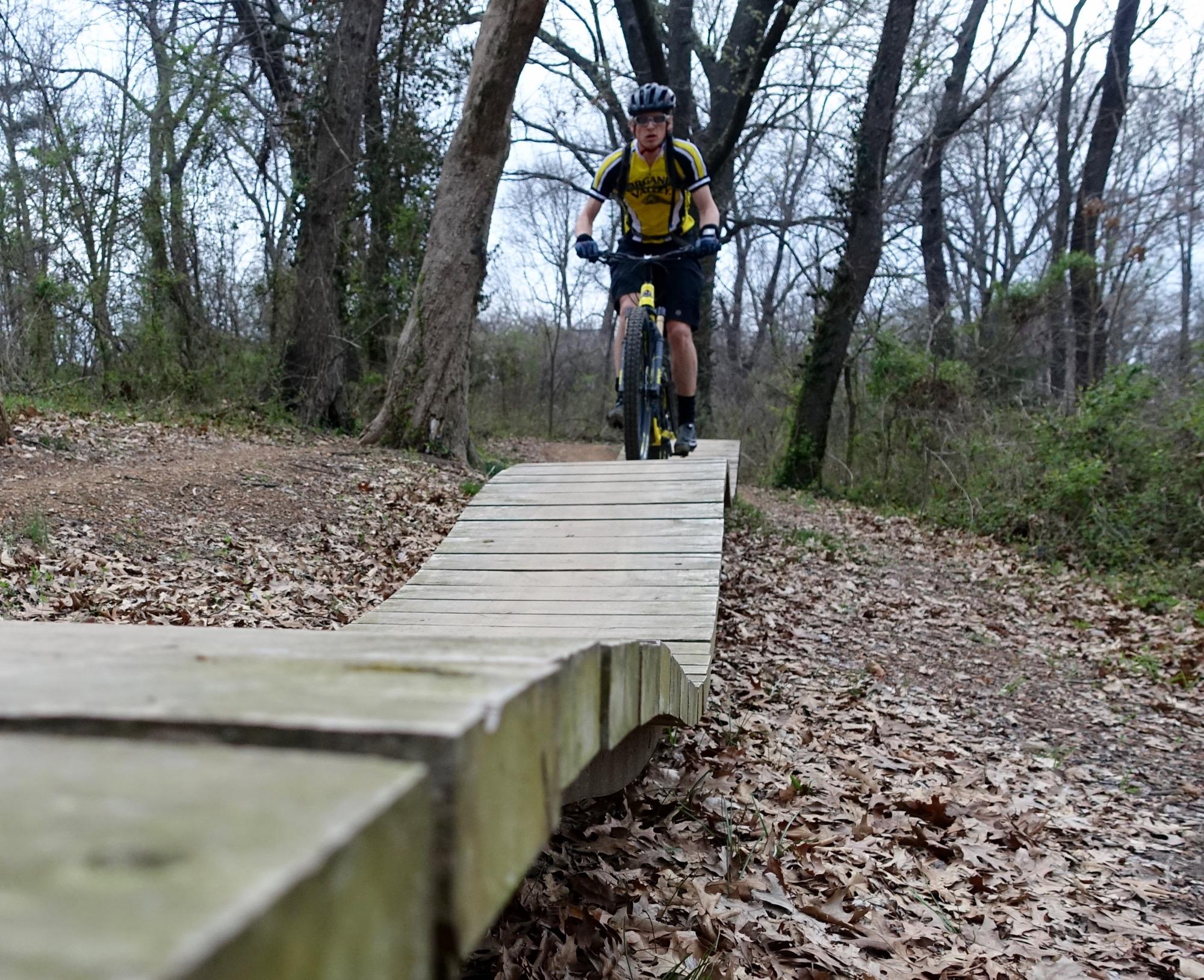 A mountain biker in a yellow jersey rides on a wooden bridge over a forest path, surrounded by trees and fallen leaves. The bridge curves as it stretches along the trail, illustrating an adventurous biking route. Slaughter Pen Trail mountain bike trail.