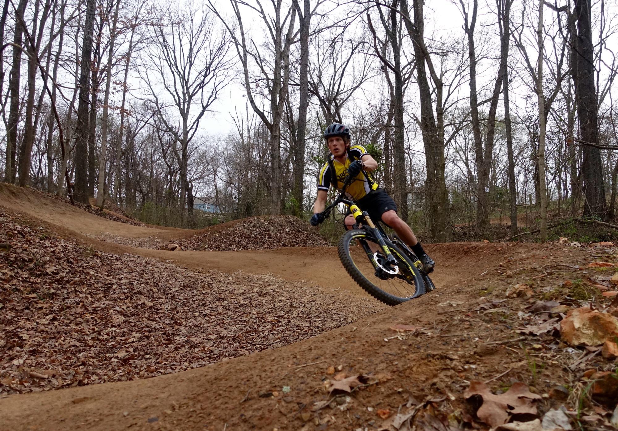 A mountain biker wearing a yellow and black jersey navigates a dirt track in a forested area, leaning into a turn with trees in the background and fallen leaves scattered on the ground. Slaughter Pen Trail mountain bike trail.