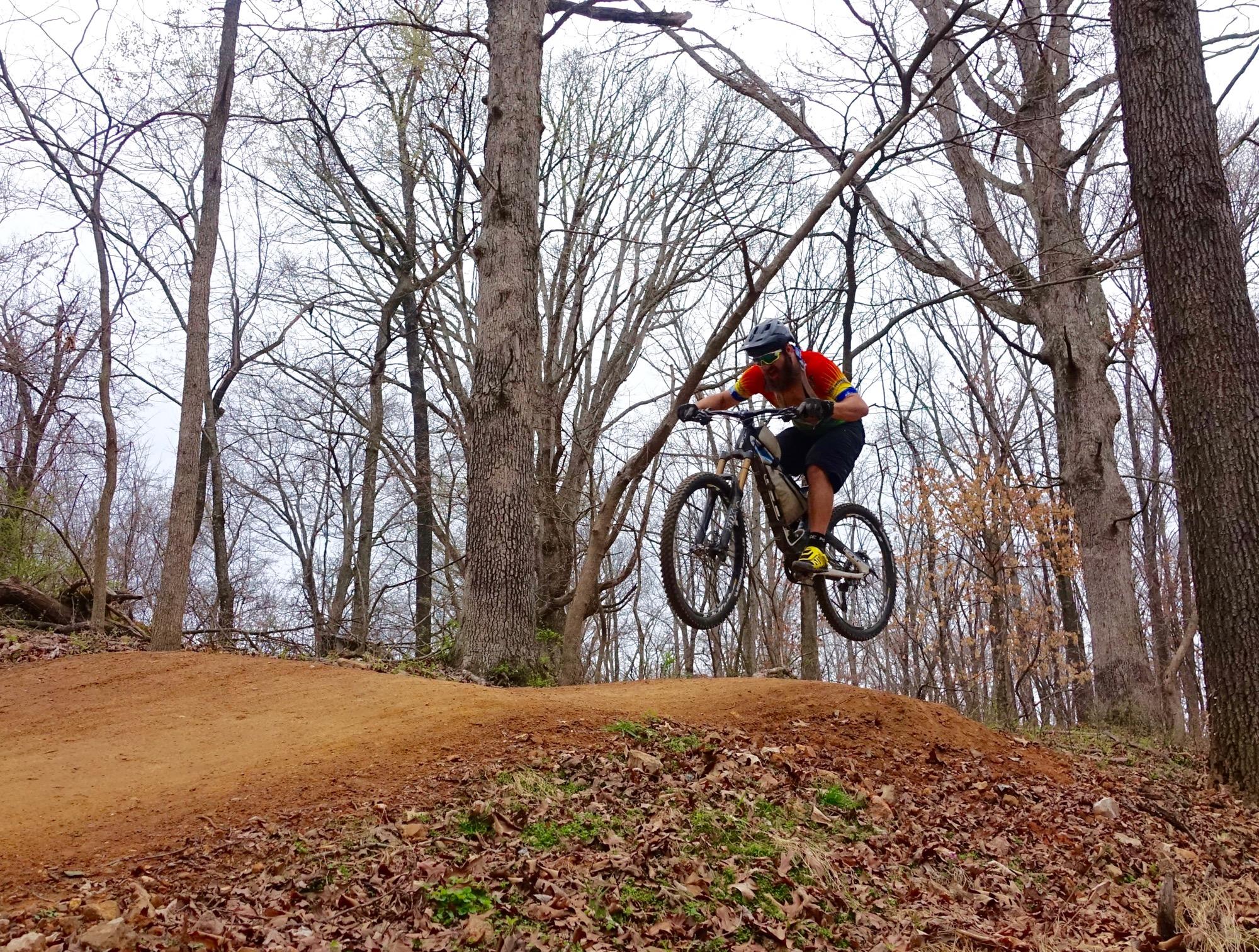 A mountain biker in mid-air, performing a jump over a dirt mound, surrounded by bare trees and autumn leaves on the ground in a forested setting. The sky is overcast, creating a cool atmosphere. The rider is wearing a helmet and colorful jersey. Slaughter Pen Trail mountain bike trail.
