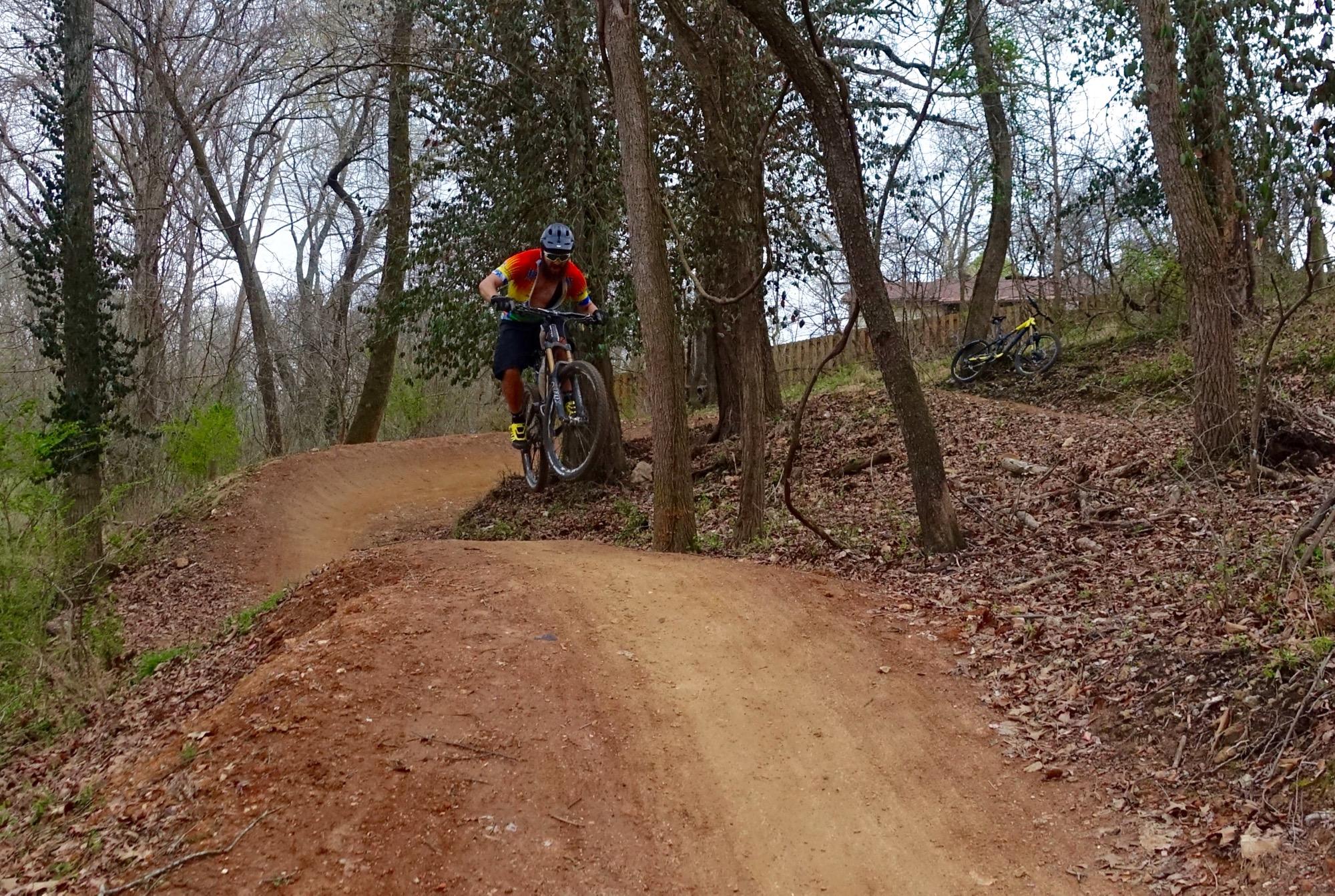 A mountain biker in a colorful jersey and helmet is airborne over a dirt jump on a forested trail, surrounded by trees and foliage. A second bike is leaned against a tree in the background. The scene captures a dynamic moment in outdoor cycling. Slaughter Pen Trail mountain bike trail.