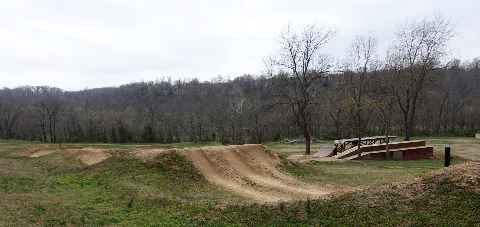 A dirt bike park featuring several dirt jumps and a wooden ramp setup, surrounded by grassy fields and trees in the background. The sky is cloudy, suggesting an overcast day. Slaughter Pen Trail mountain bike trail.