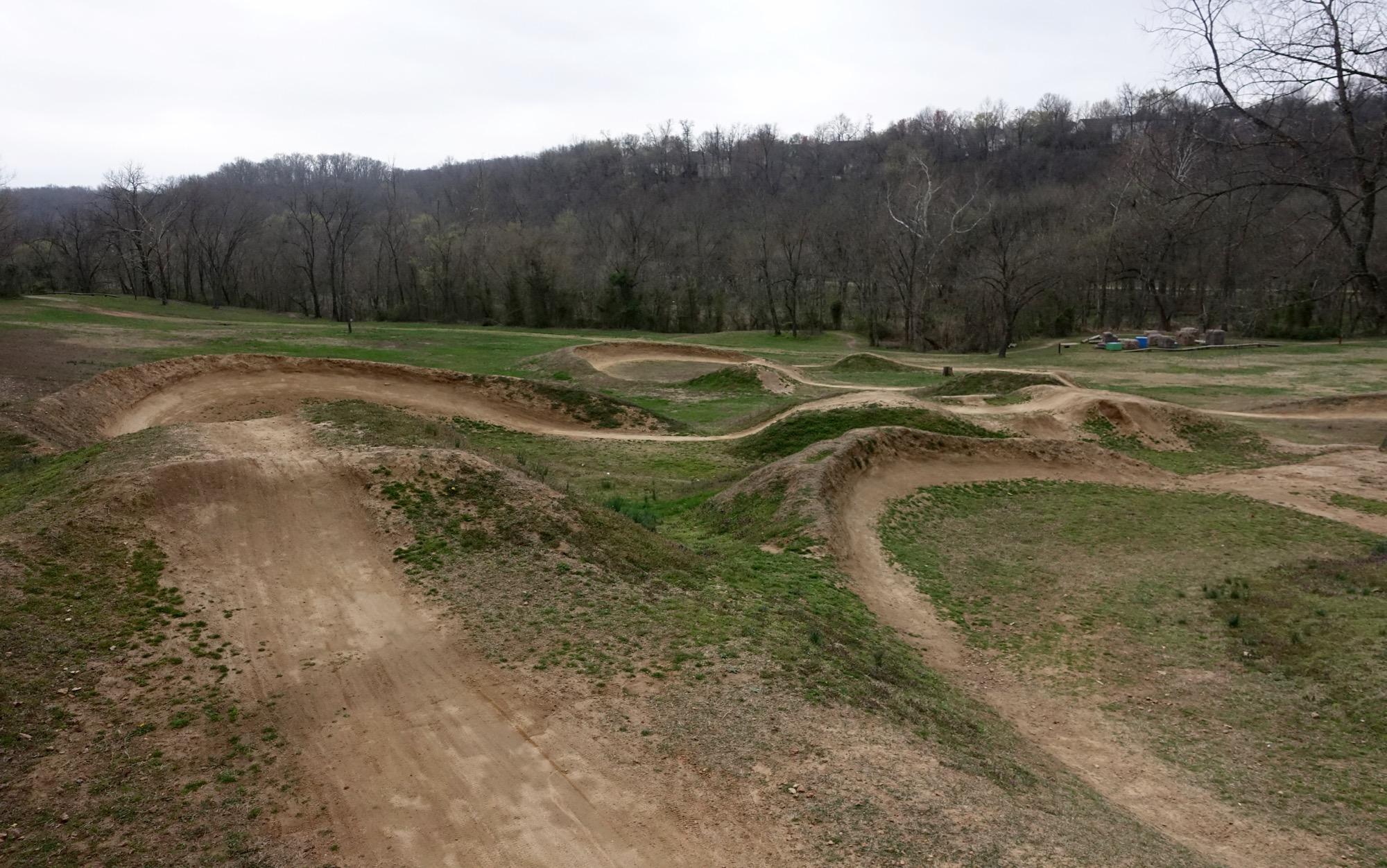 A dirt bike track featuring a series of hilly curves and jumps, surrounded by grassy areas and sparse trees, under a cloudy sky. Slaughter Pen Trail mountain bike trail.