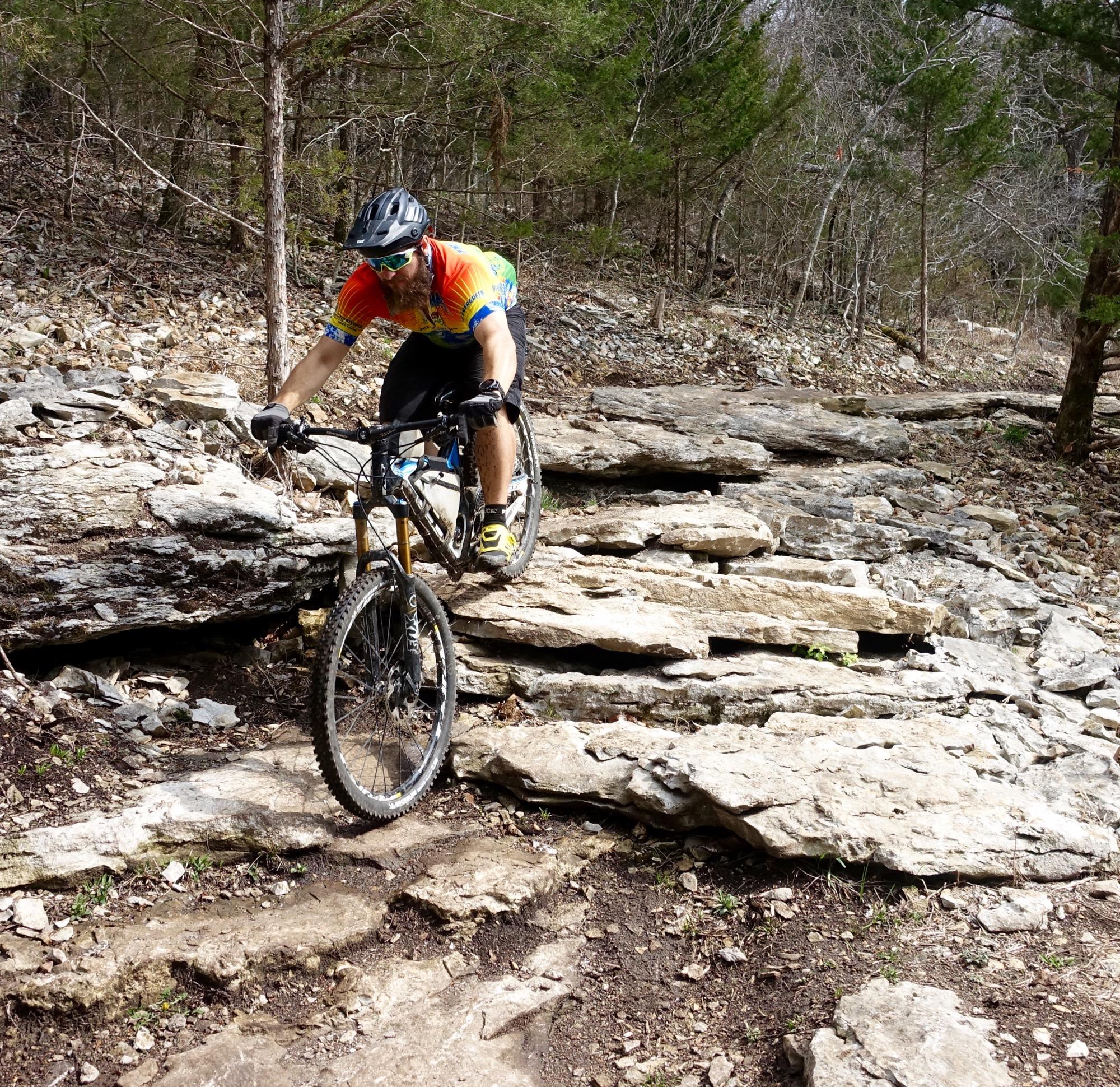 A mountain biker navigating a rocky trail, showcasing balance and skill as he rides over large stones in a forested area. Coler Mountain Bike Preserve mountain bike trail.