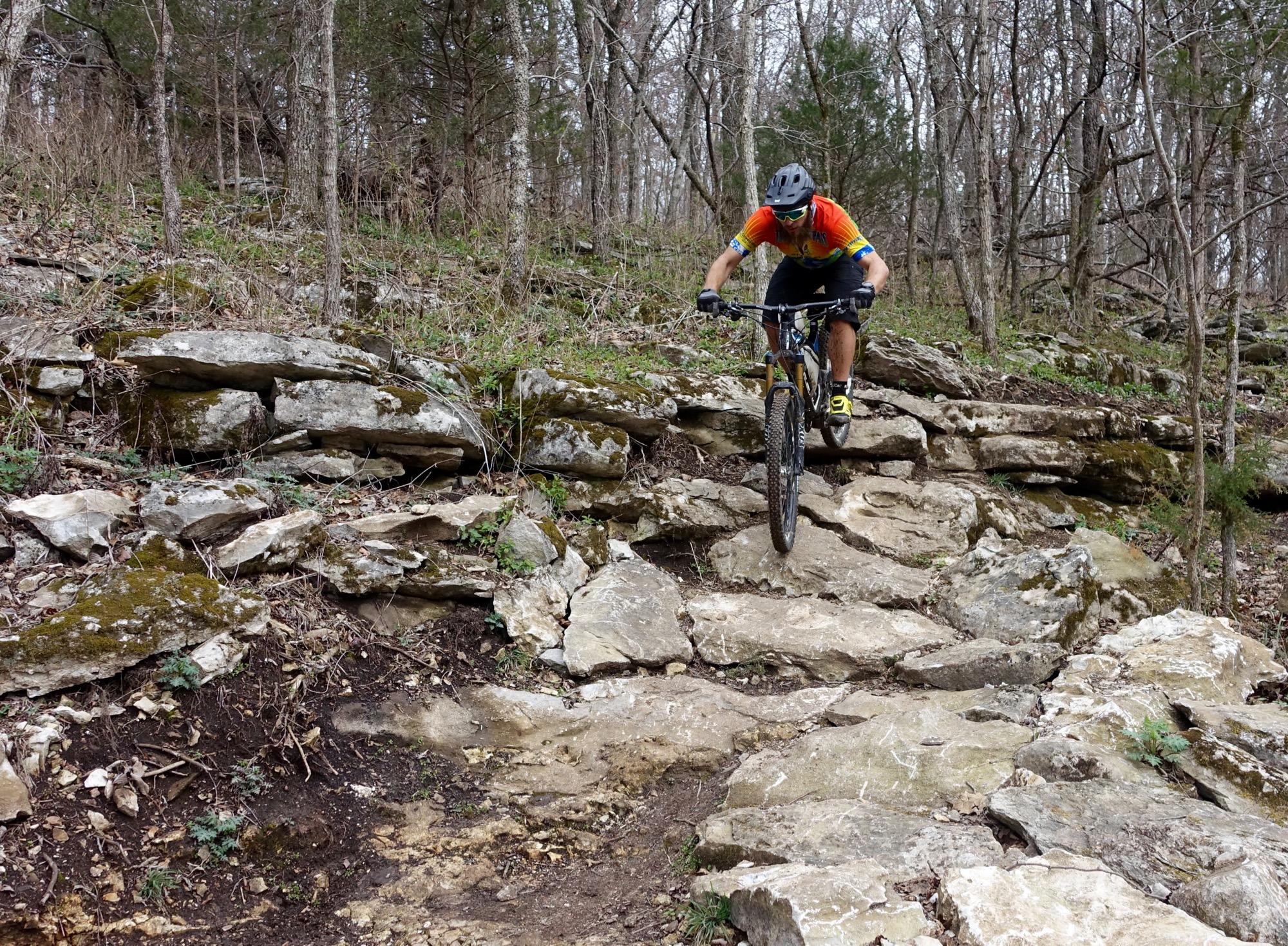 A mountain biker navigating rocky terrain in a forested area, wearing a helmet and brightly colored jersey. The bike is mid-motion, showcasing a challenging descent over uneven rocks and sparse vegetation. Coler Mountain Bike Preserve mountain bike trail.