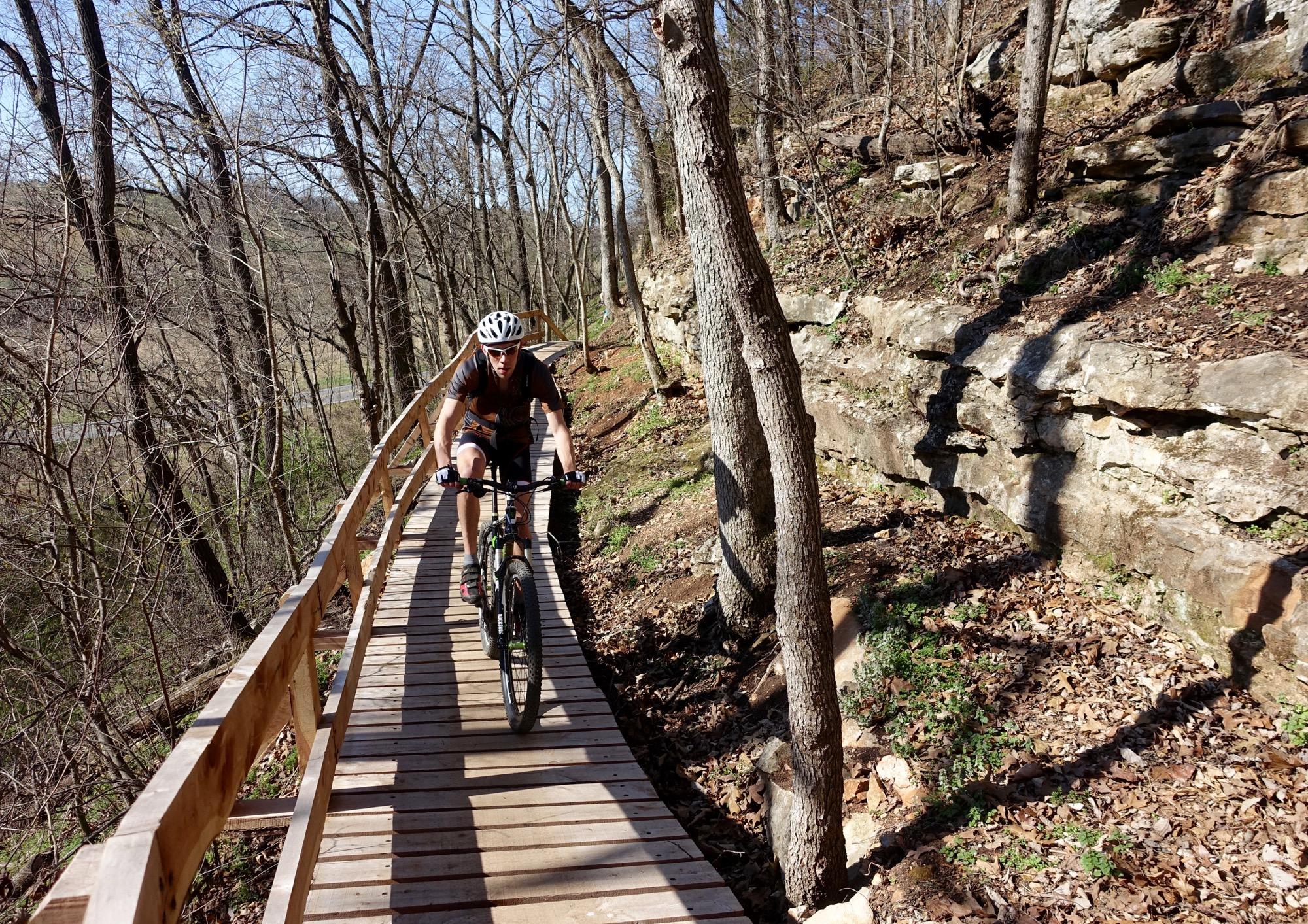 A cyclist riding a mountain bike along a wooden path in a wooded area, surrounded by trees and rocky terrain, on a sunny day. Back 40 mountain bike trail.
