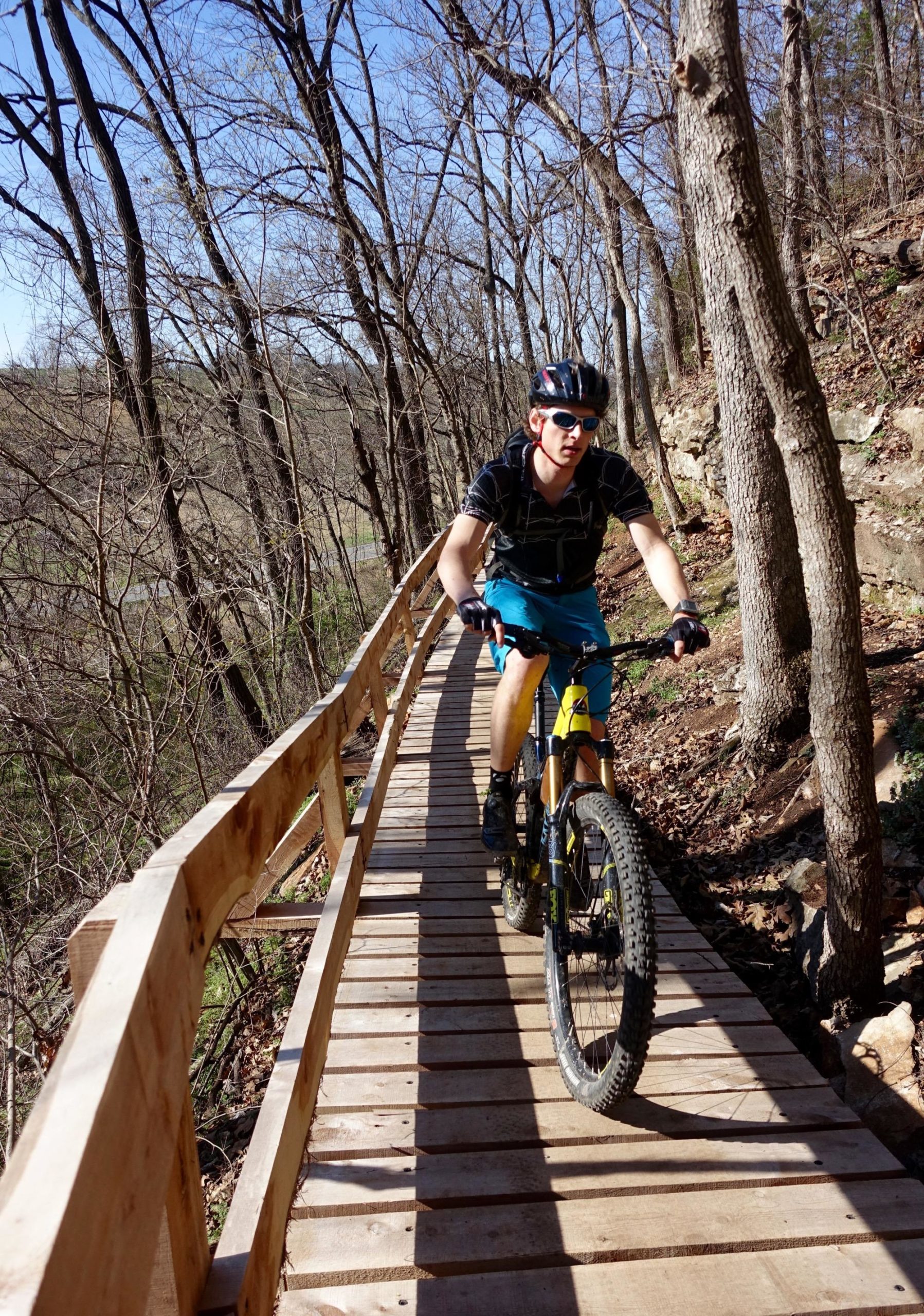 A mountain biker in a black shirt and blue shorts rides a yellow bike along a wooden path in a forested area, surrounded by bare trees and a clear blue sky. Back 40 mountain bike trail.